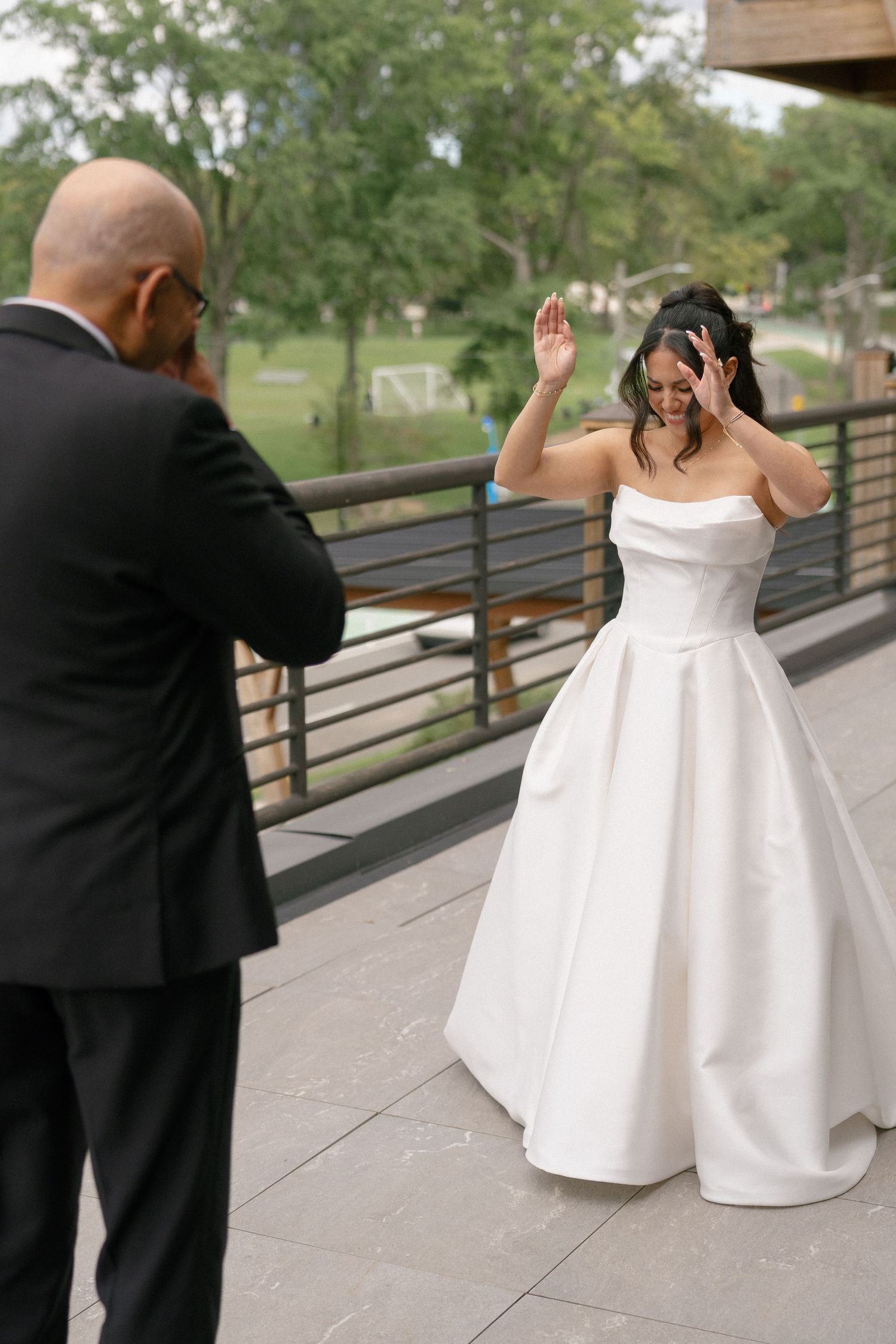 Bride in white strapless ball gown waves joyfully during first look moment with groom in black suit on outdoor terrace.