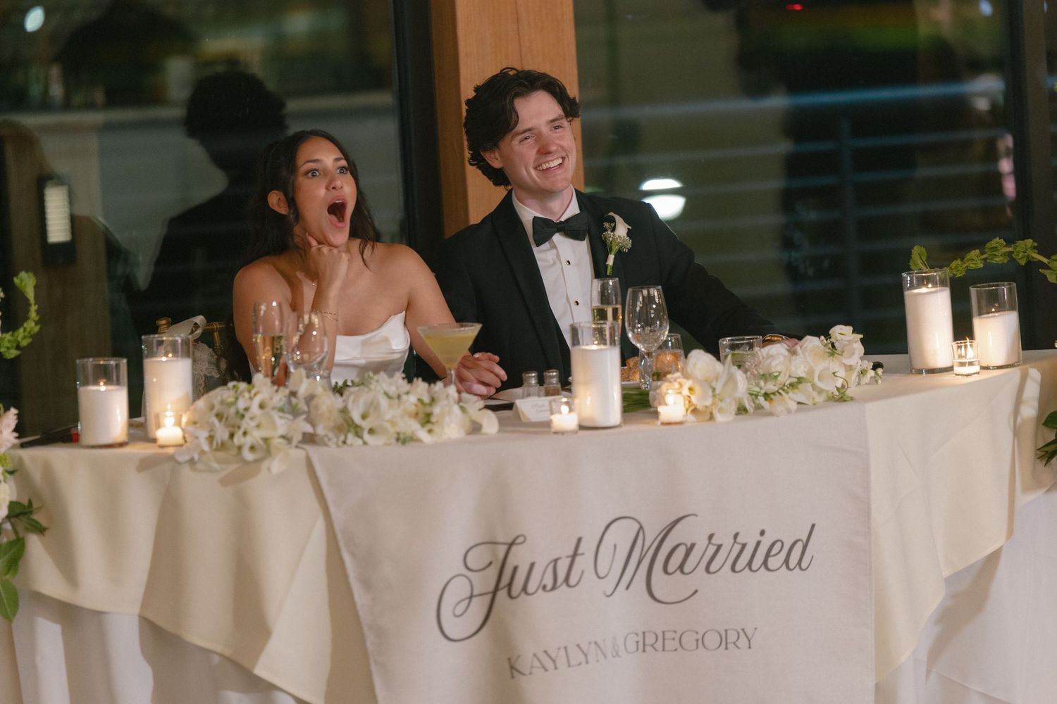 Bride and groom laughing at Just Married sweetheart table decorated with white florals and candles at elegant wedding reception.