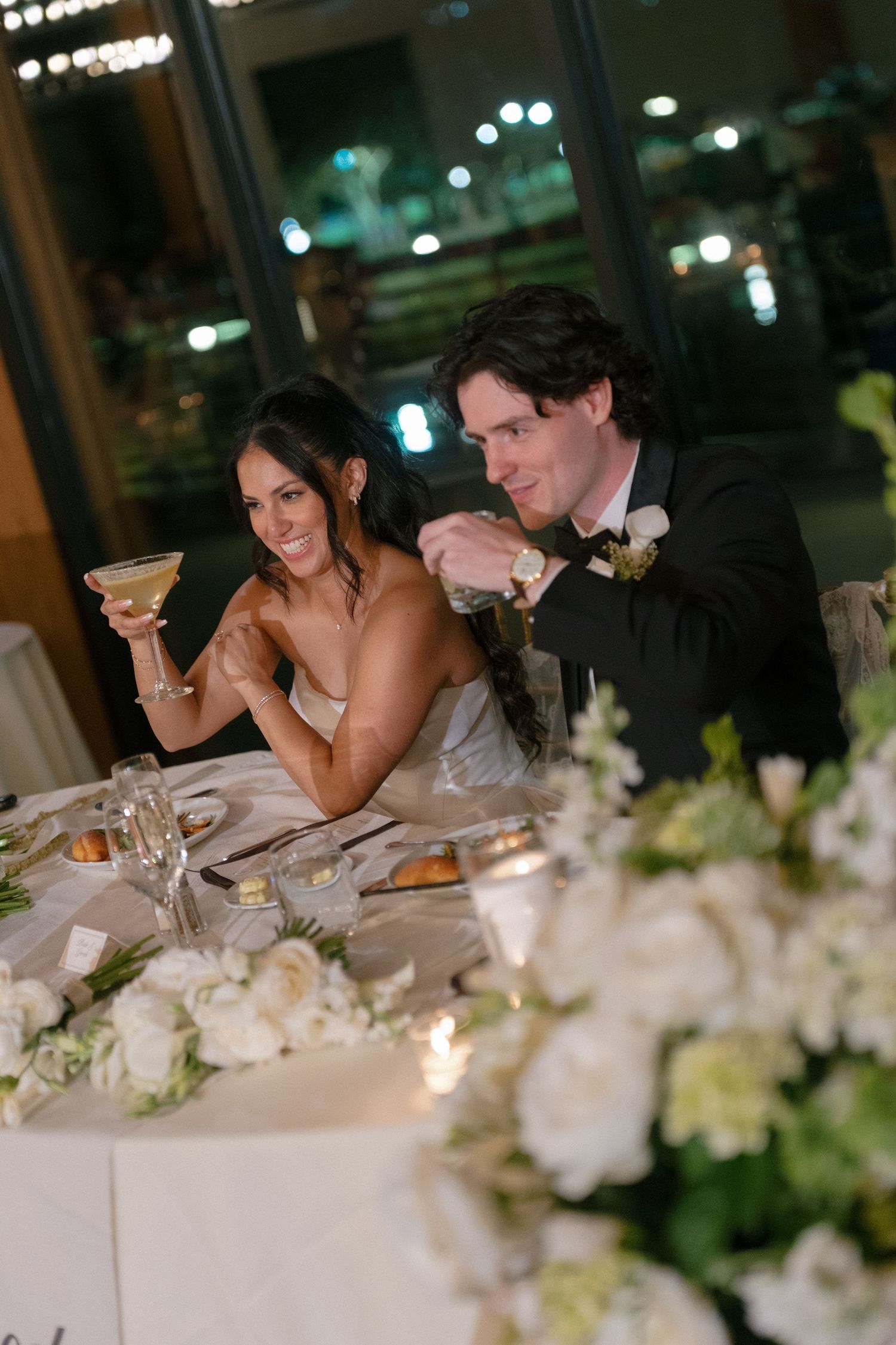 Bride and groom toast at sweetheart table adorned with white floral arrangements during wedding reception.