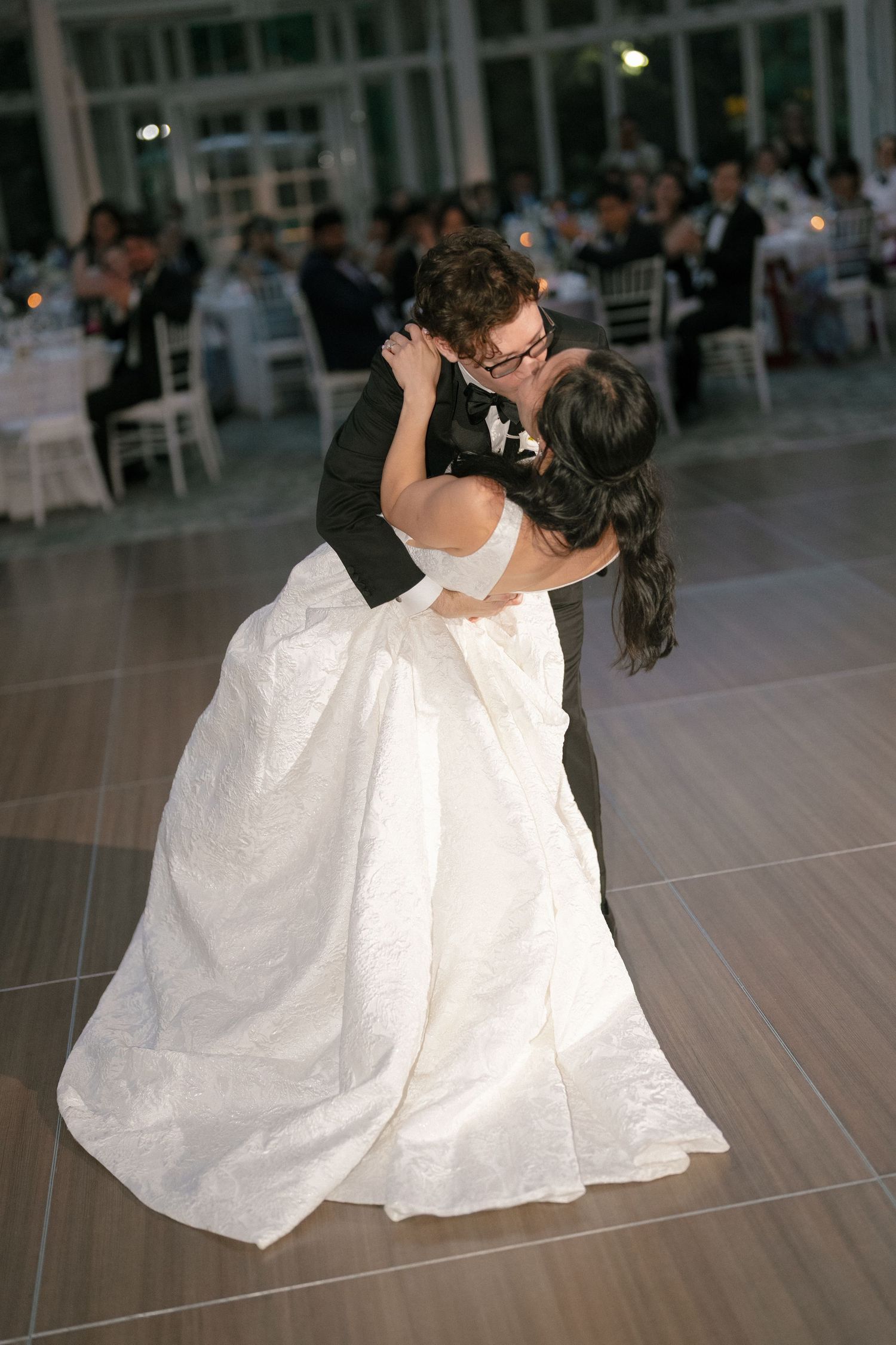 Bride and groom share a romantic dip kiss during their first dance at an elegant indoor wedding reception.