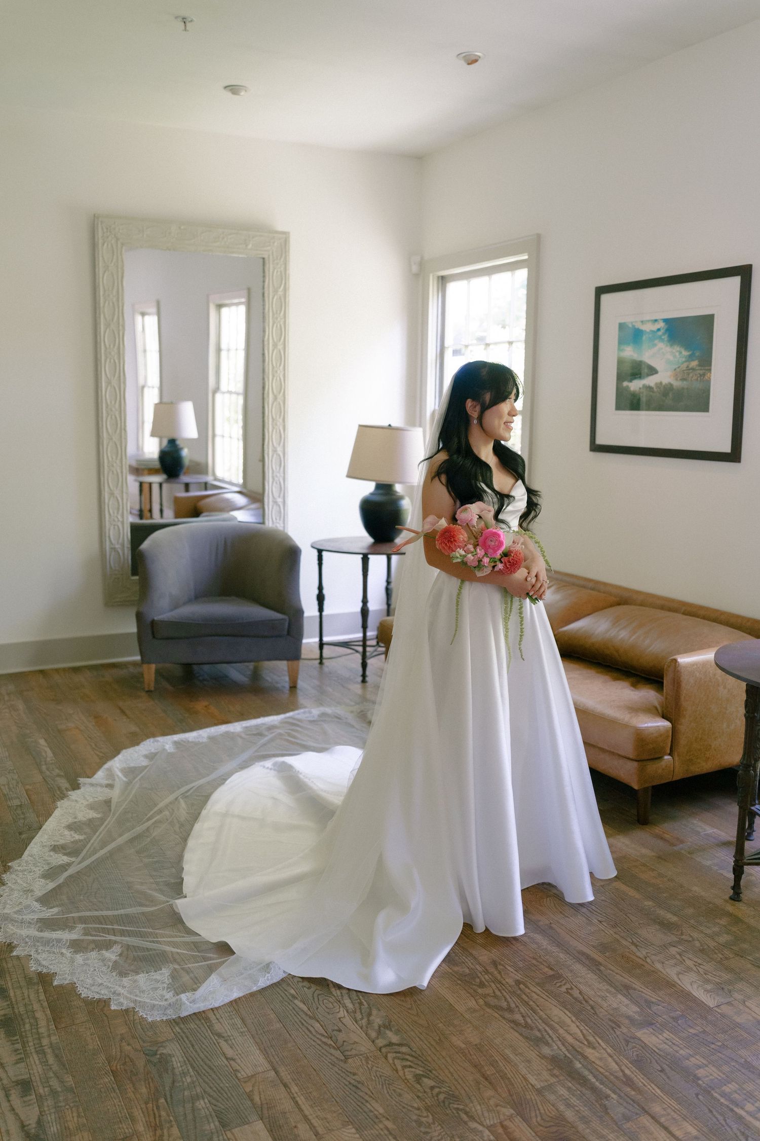 Bride in a white strapless gown with cathedral veil holds pink bouquet in an elegant room.