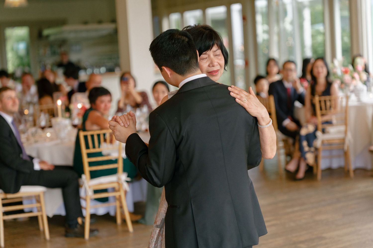 Groom dancing with his mother at a wedding reception while guests applaud and watch in a bright elegant venue.