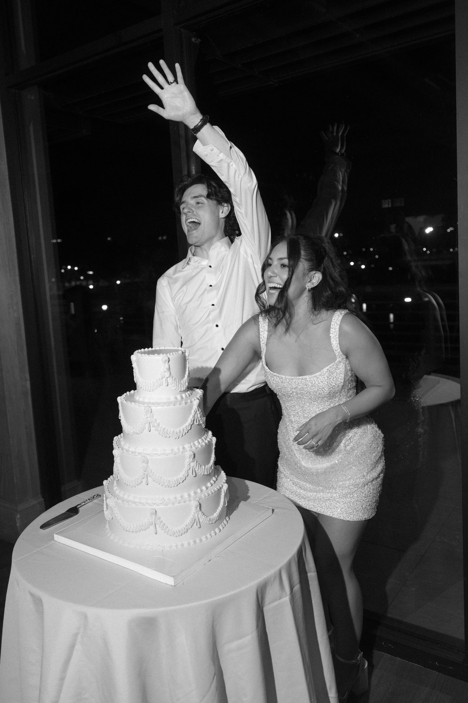Black and white photo of excited bride and groom celebrating wedding cake cutting with hands raised in joy.