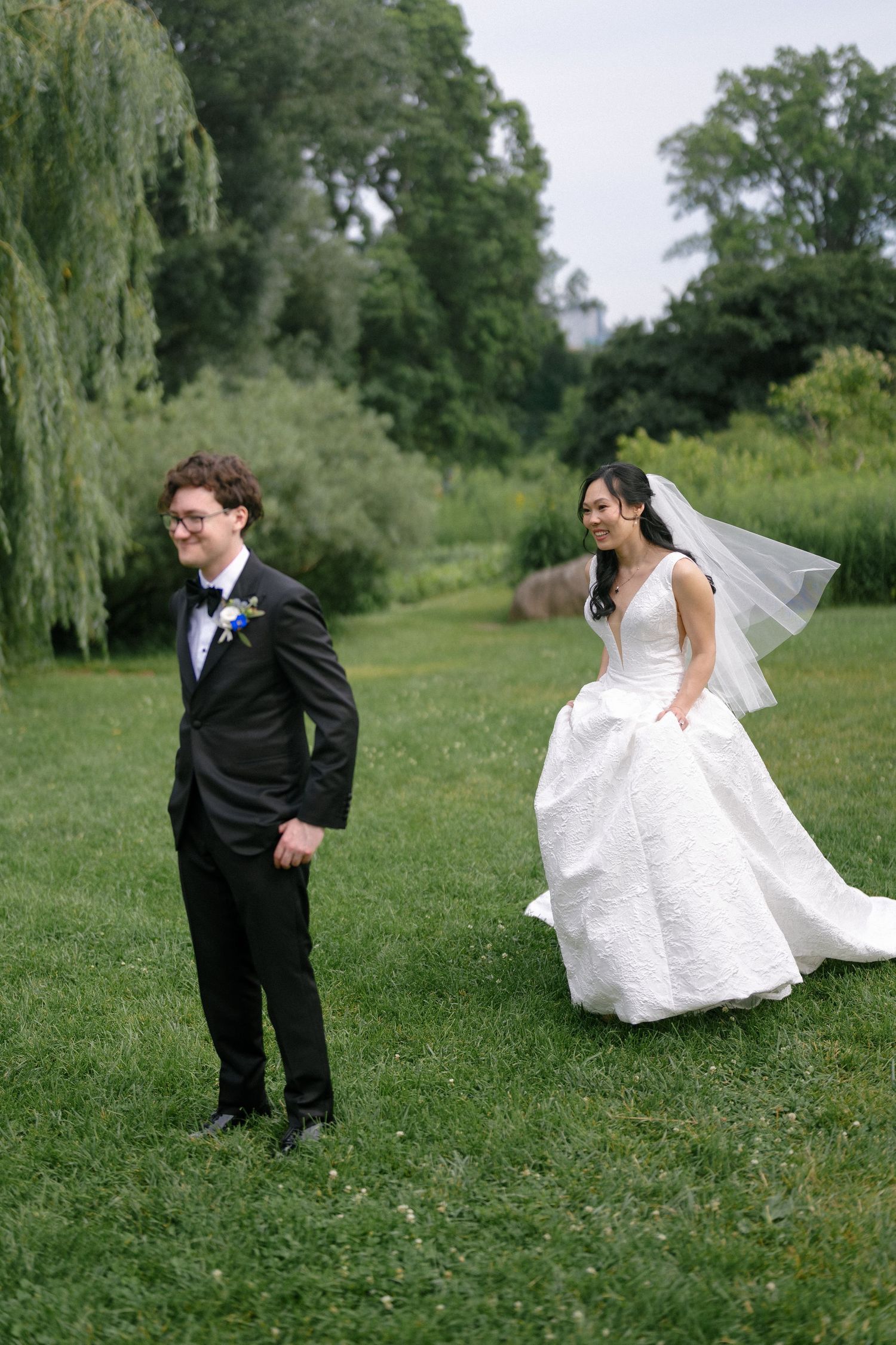 Bride in white ball gown with veil walks toward groom in tuxedo on lush green lawn at outdoor wedding.