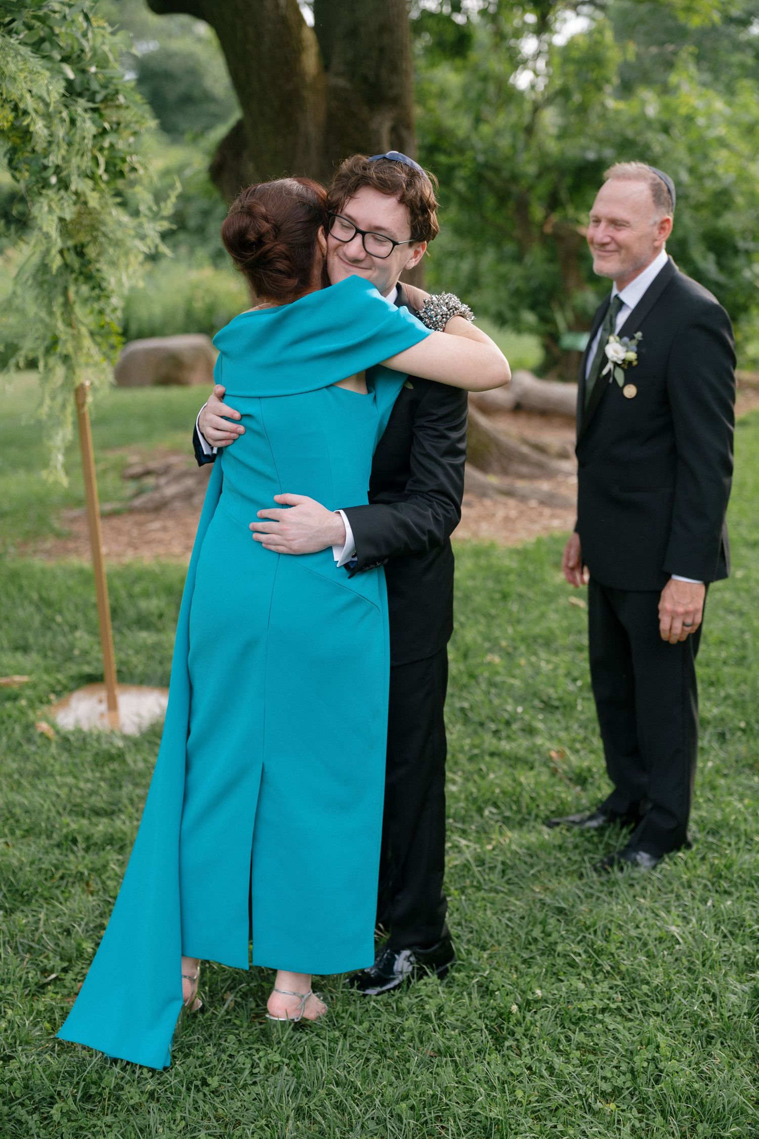 Person in teal dress hugging someone in black suit outdoors at a wedding ceremony with a smiling man in background.