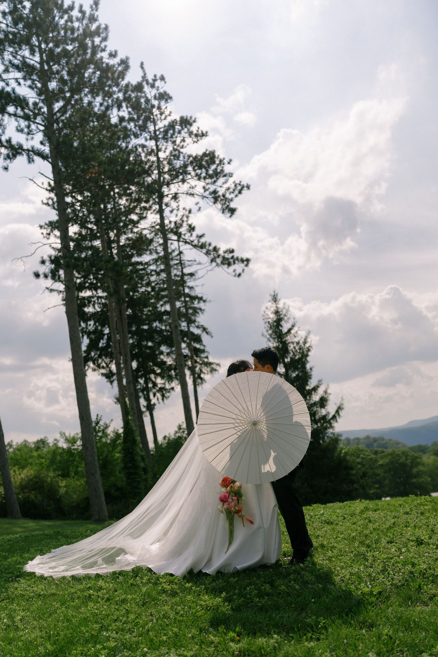 Bride and groom share a romantic moment outdoors, bride holding a white parasol and bouquet, with tall pine trees and cloudy sky.
