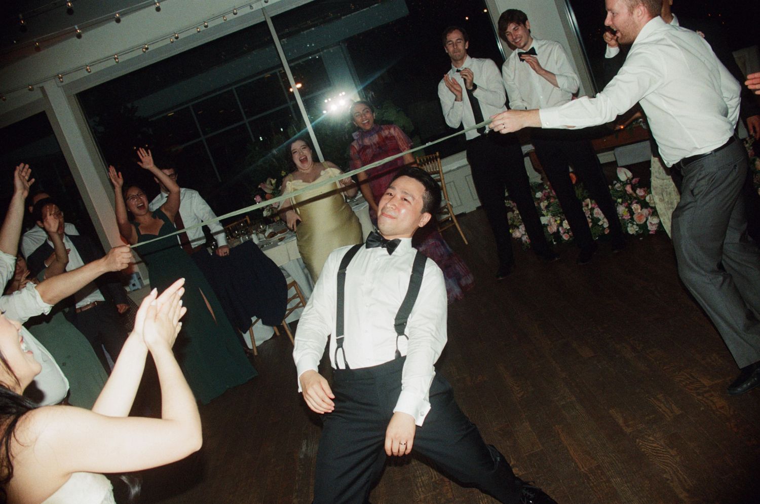 Groom in suspenders dancing energetically on wedding reception dance floor surrounded by guests.