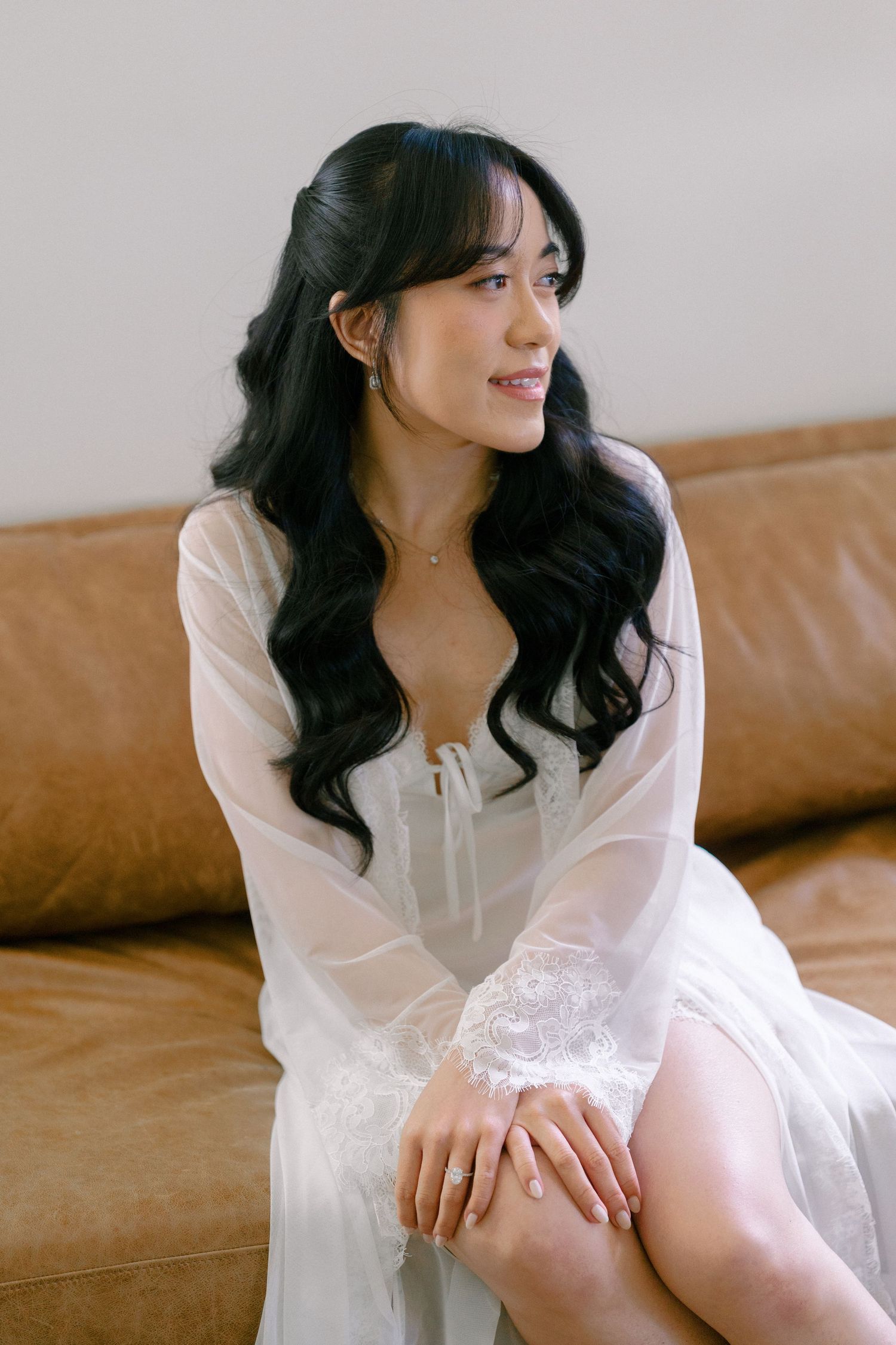 Bride in white lace robe sitting on brown leather sofa with wavy black hair and delicate jewelry.