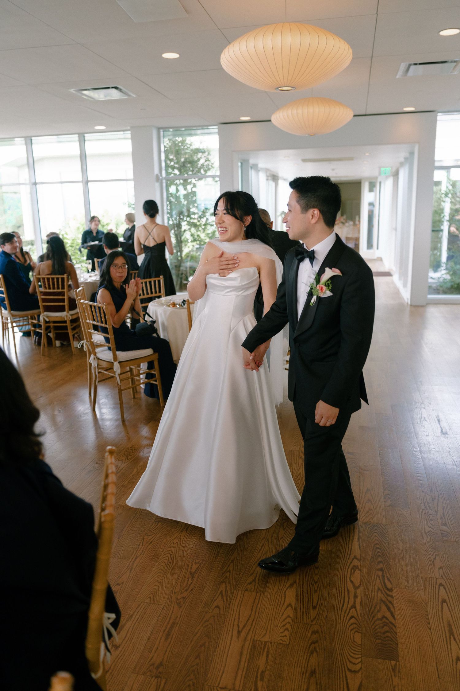 Bride and groom making grand entrance into bright wedding reception hall with gold chiavari chairs and guests seated.