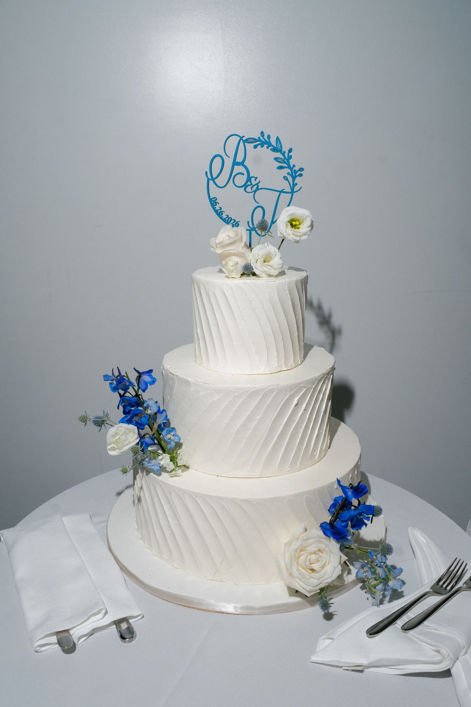 Elegant three-tier white wedding cake with blue floral decorations and monogram topper on white tablecloth.
