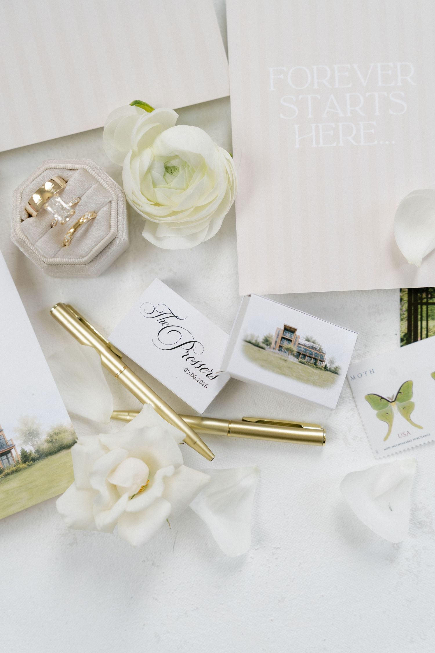 Wedding flat lay with white ranunculus flowers, gold pens, ring box, and Forever Starts Here sign.