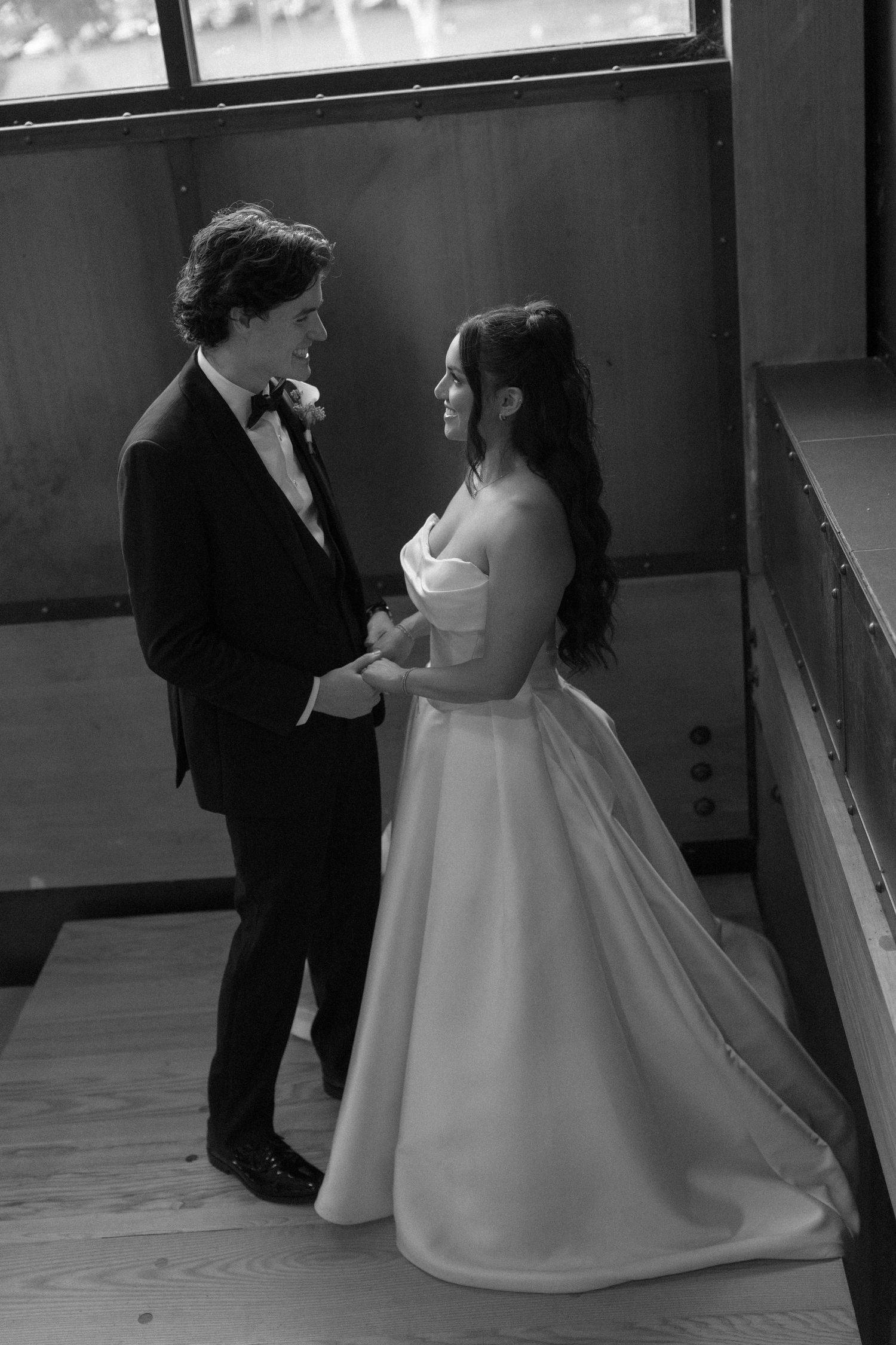 Black and white photo of bride and groom holding hands smiling at each other on a staircase indoors.