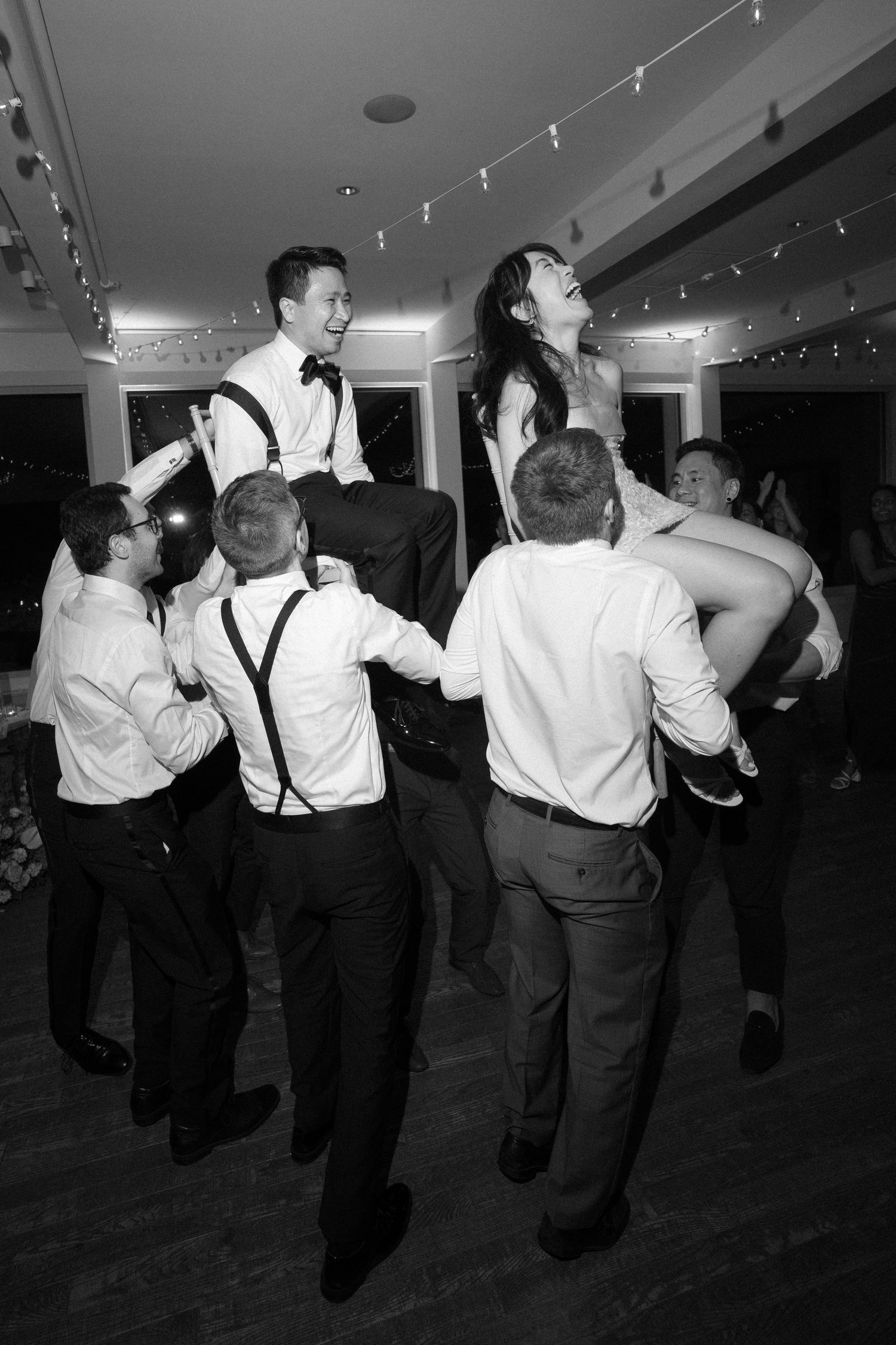 Black and white photo of wedding guests lifting bride and groom during lively reception dance.