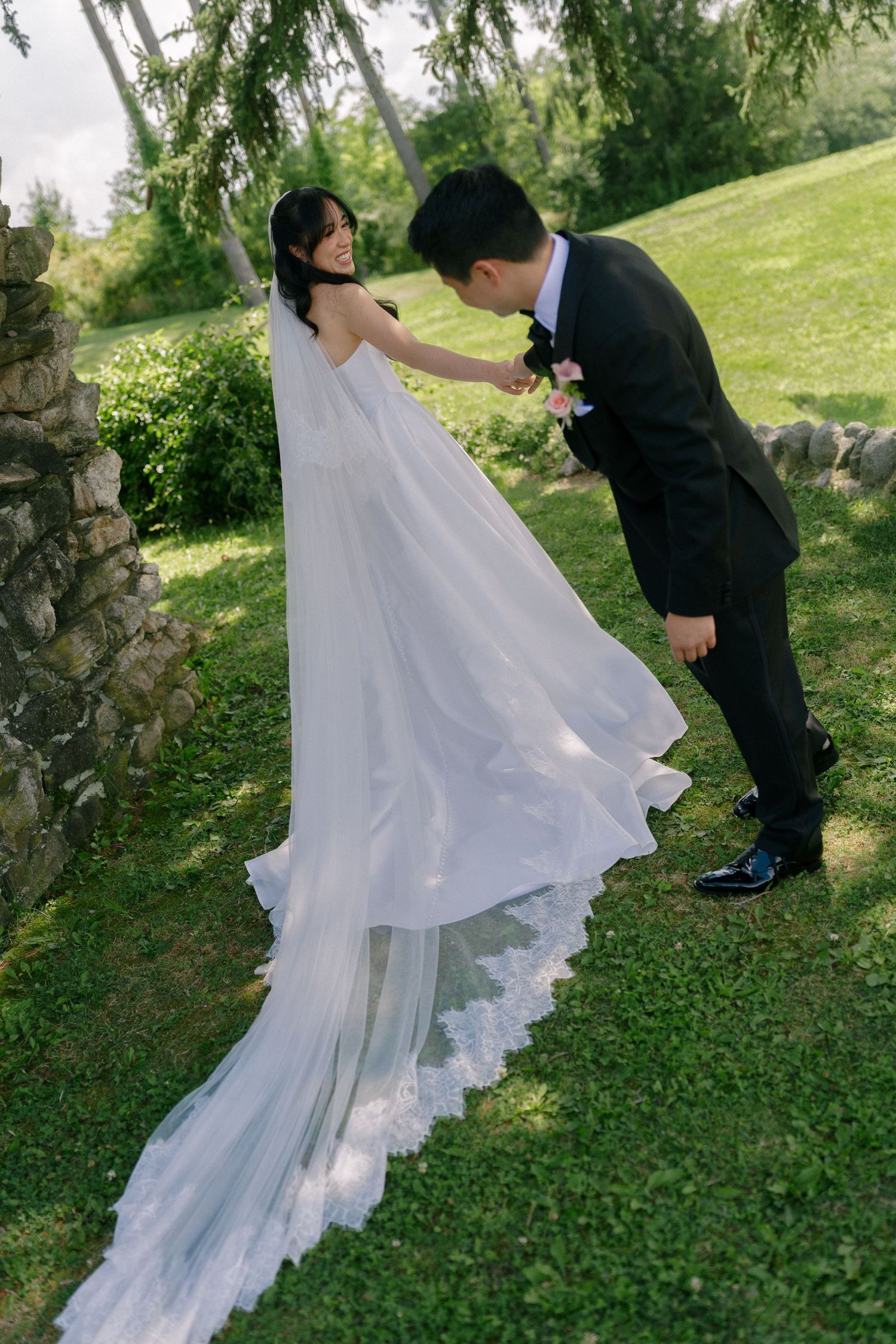 Groom bows to bride in white gown with lace cathedral veil in a lush green garden.