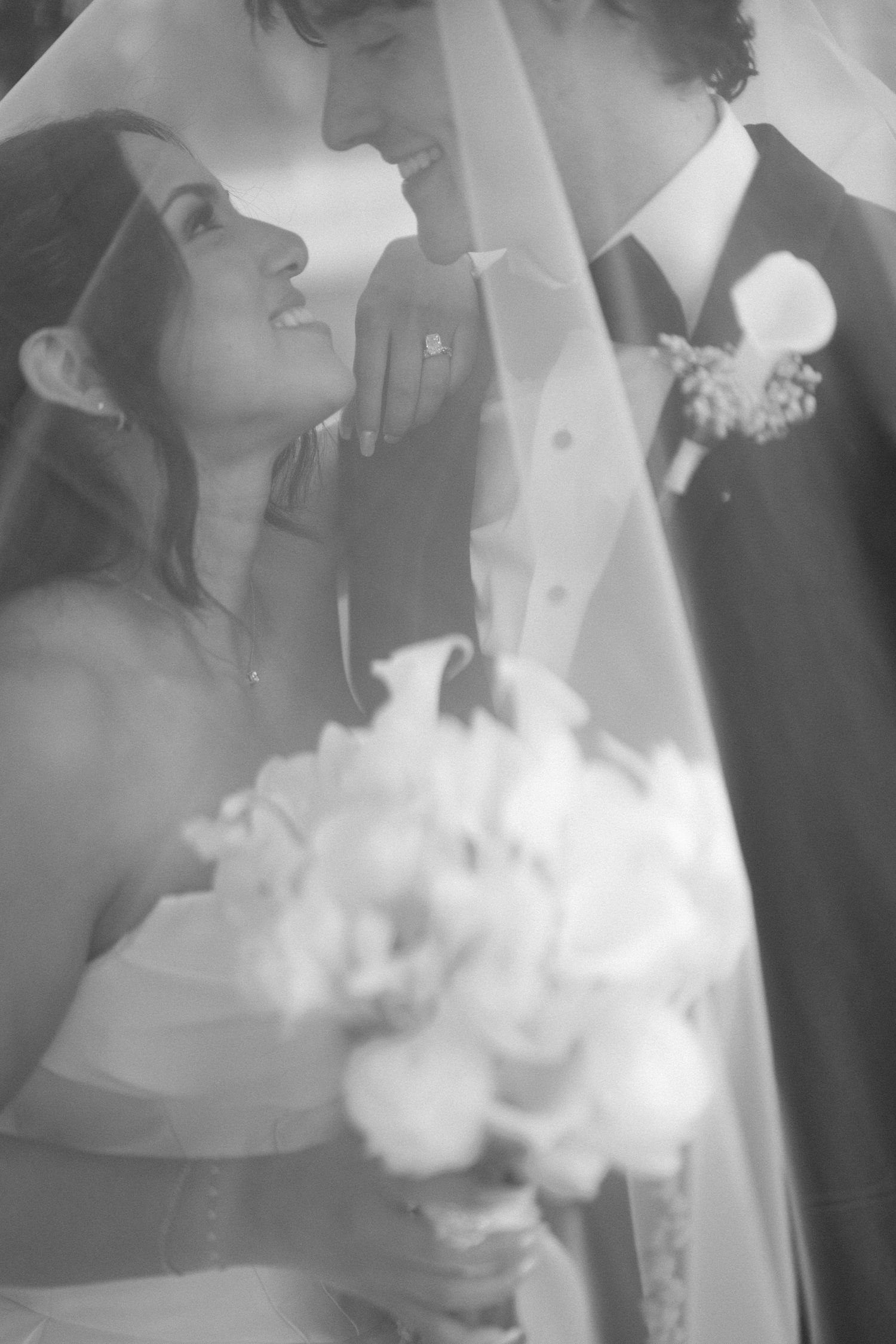 Black and white photo of bride gazing up at groom under veil, holding a white floral bouquet.