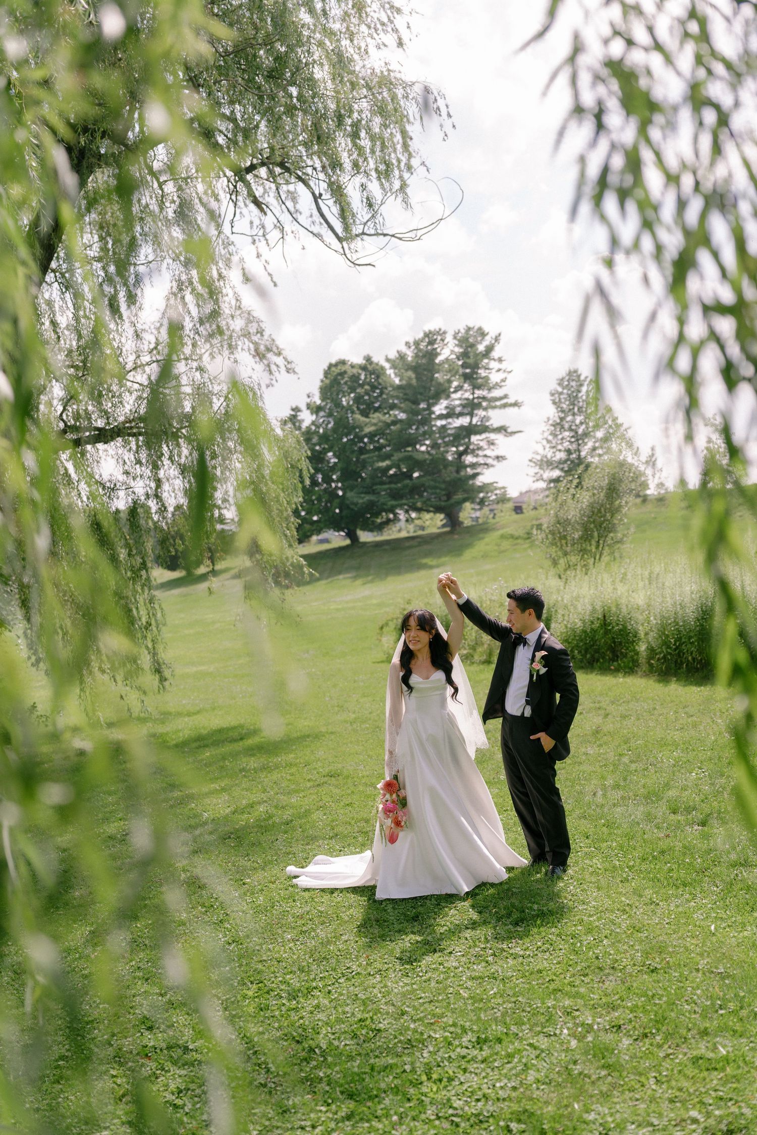 Bride and groom dancing under a weeping willow tree on a lush green lawn at an outdoor wedding venue.