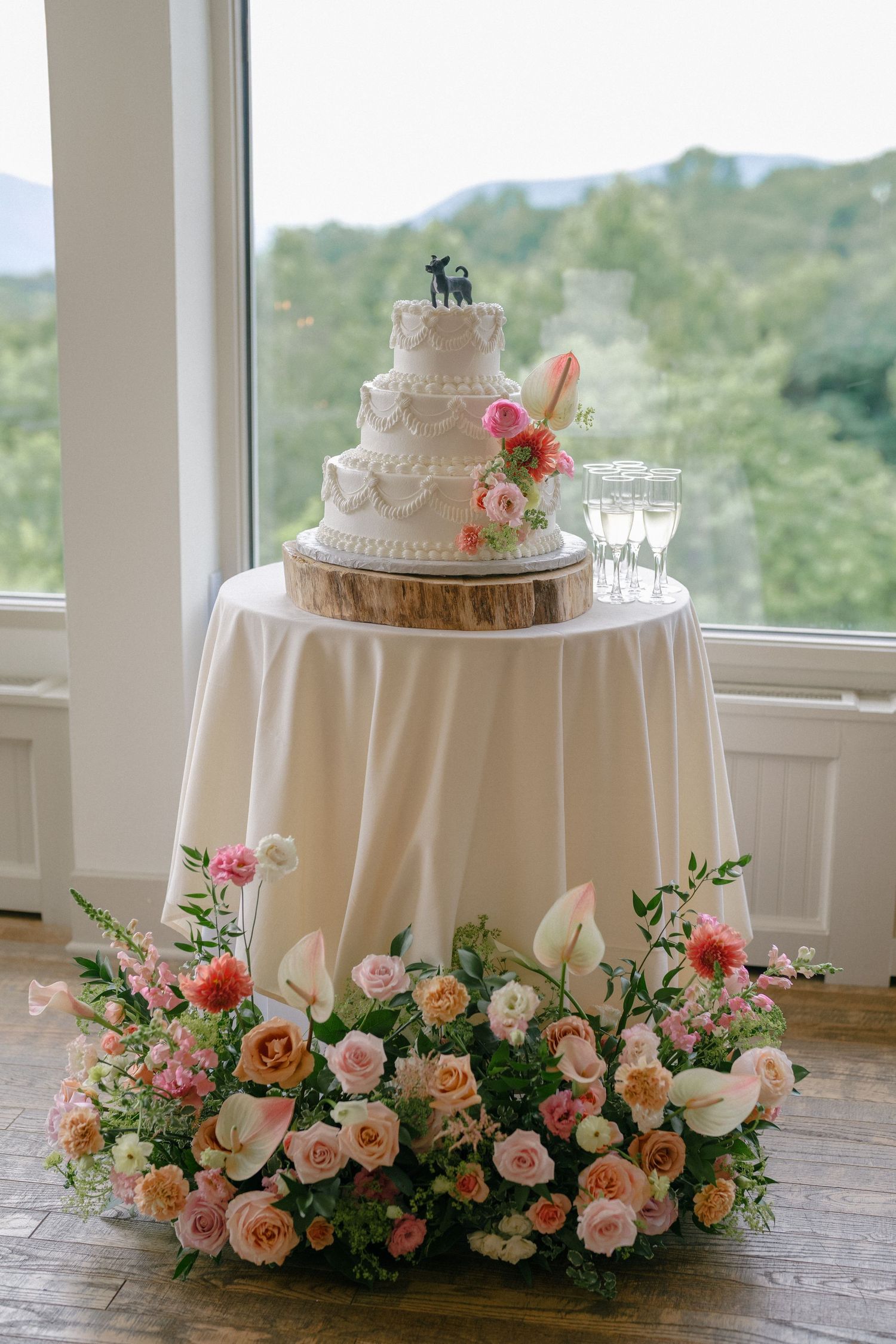 Elegant four-tier white wedding cake adorned with pink florals on a flower-surrounded table.