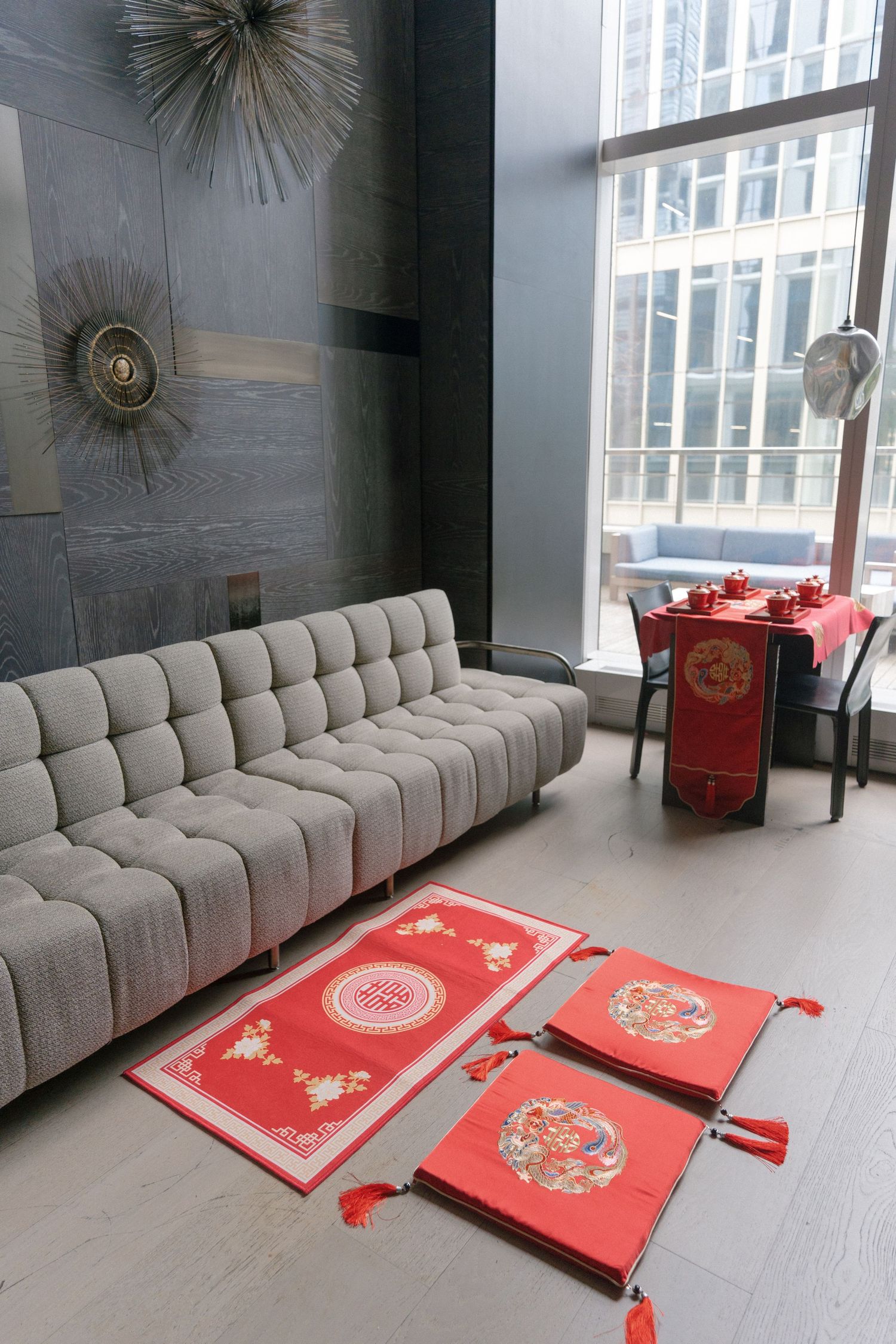 Red Chinese ceremonial table runners with tassels displayed on floor beside gray tufted sofa in modern urban venue.