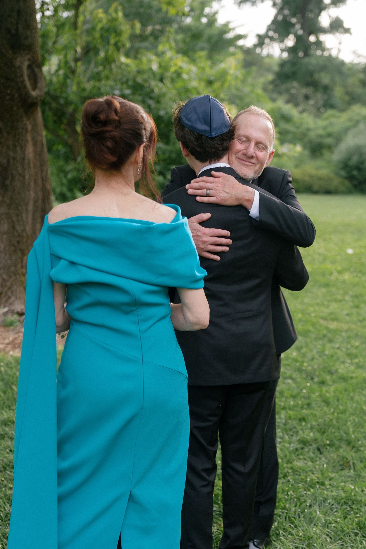 Older man in suit hugging young man in kippah while woman in teal off-shoulder gown watches at outdoor wedding.