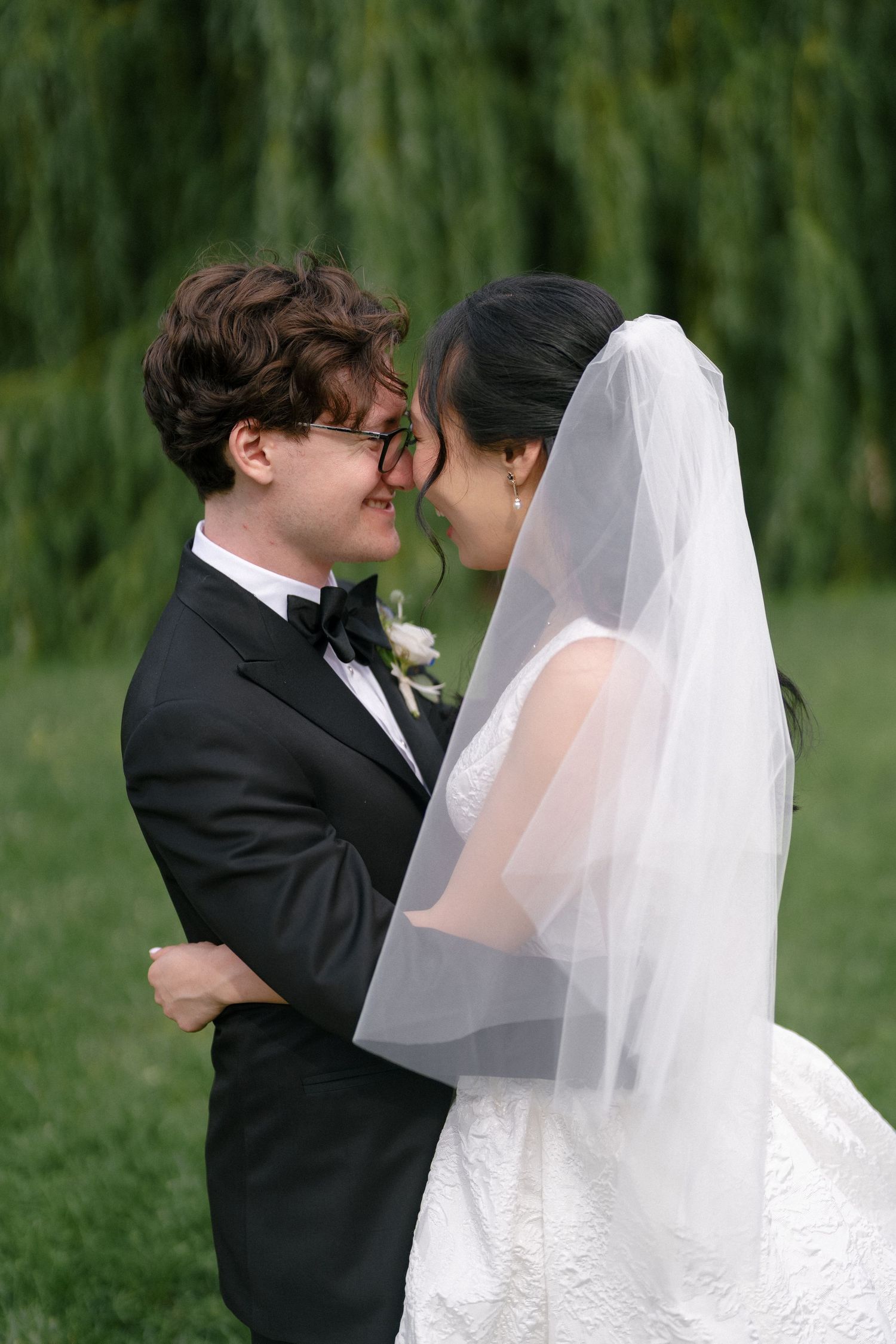 Bride and groom share romantic moment under weeping willow tree, groom in tuxedo, bride in white lace gown with veil.