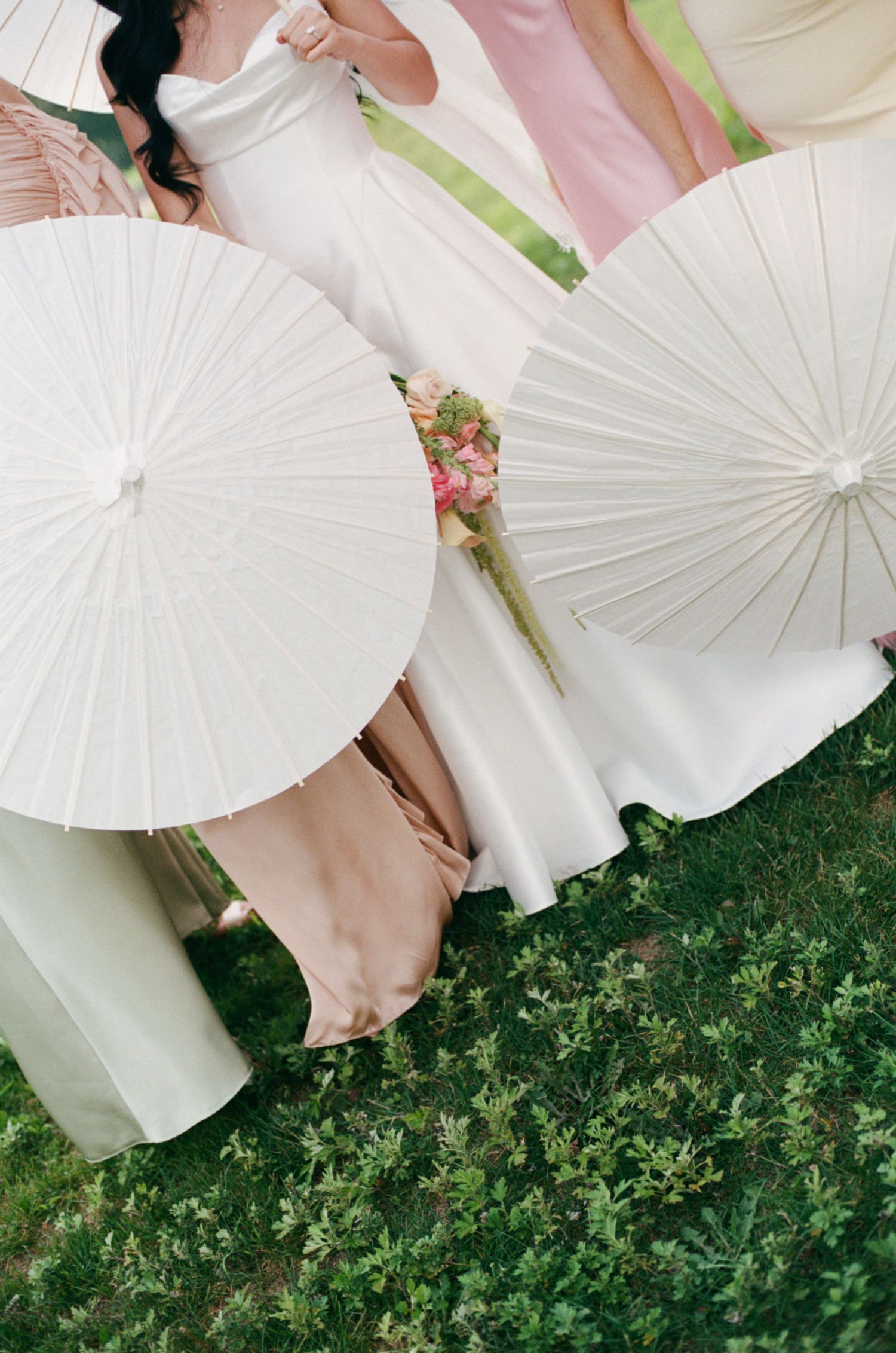 Aerial view of white paper parasols held by bride and bridesmaids in blush gowns on green lawn.