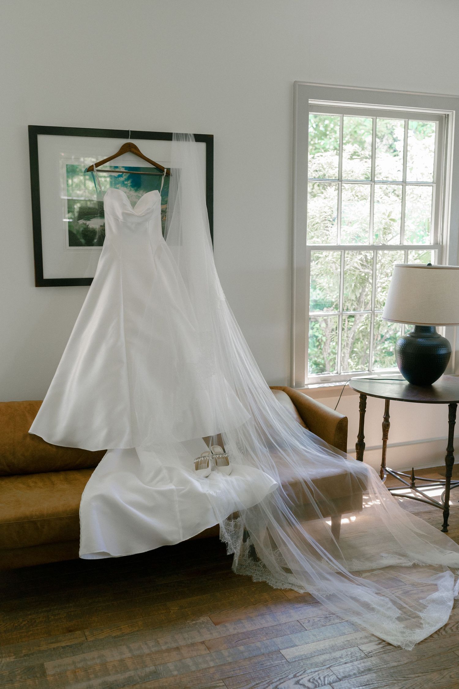 Elegant white strapless wedding gown with long veil displayed on hanger over a couch near a sunlit window.