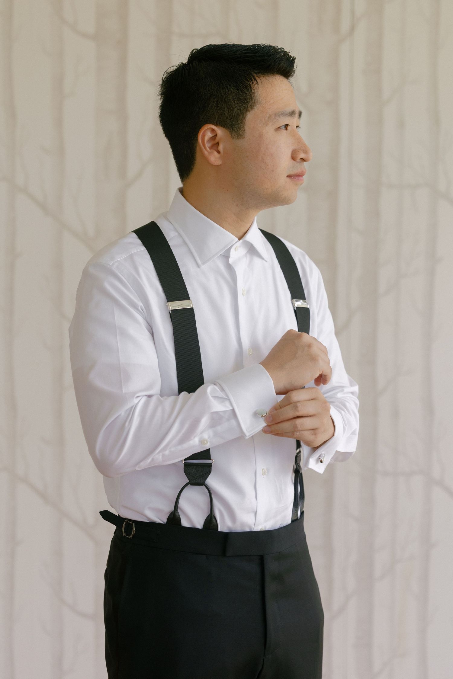 Groom in white dress shirt with black suspenders adjusting cufflinks before wedding ceremony.