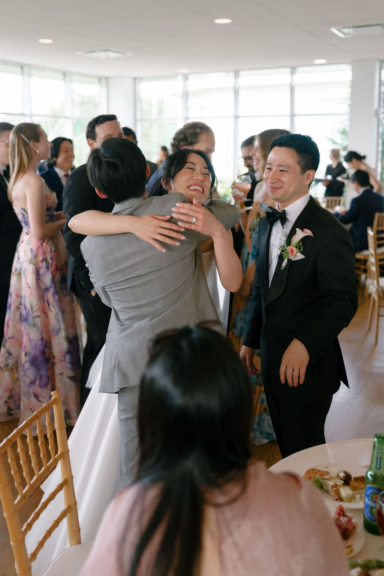 Bride laughing while hugging guest at wedding reception as groom smiles warmly beside her.