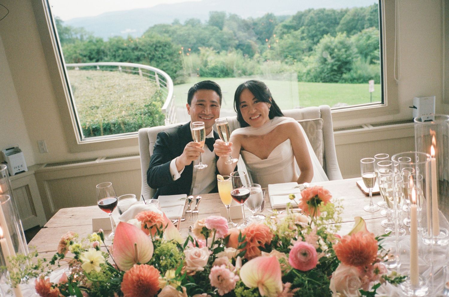 Newlyweds toast champagne at sweetheart table with pink floral centerpiece and mountain view backdrop.