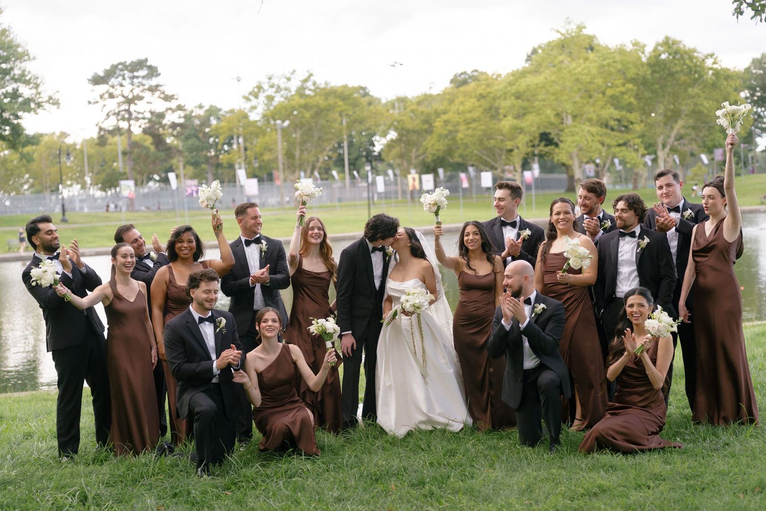 Wedding party in brown dresses and black tuxedos posing outdoors near a pond, bride and groom kissing in center.