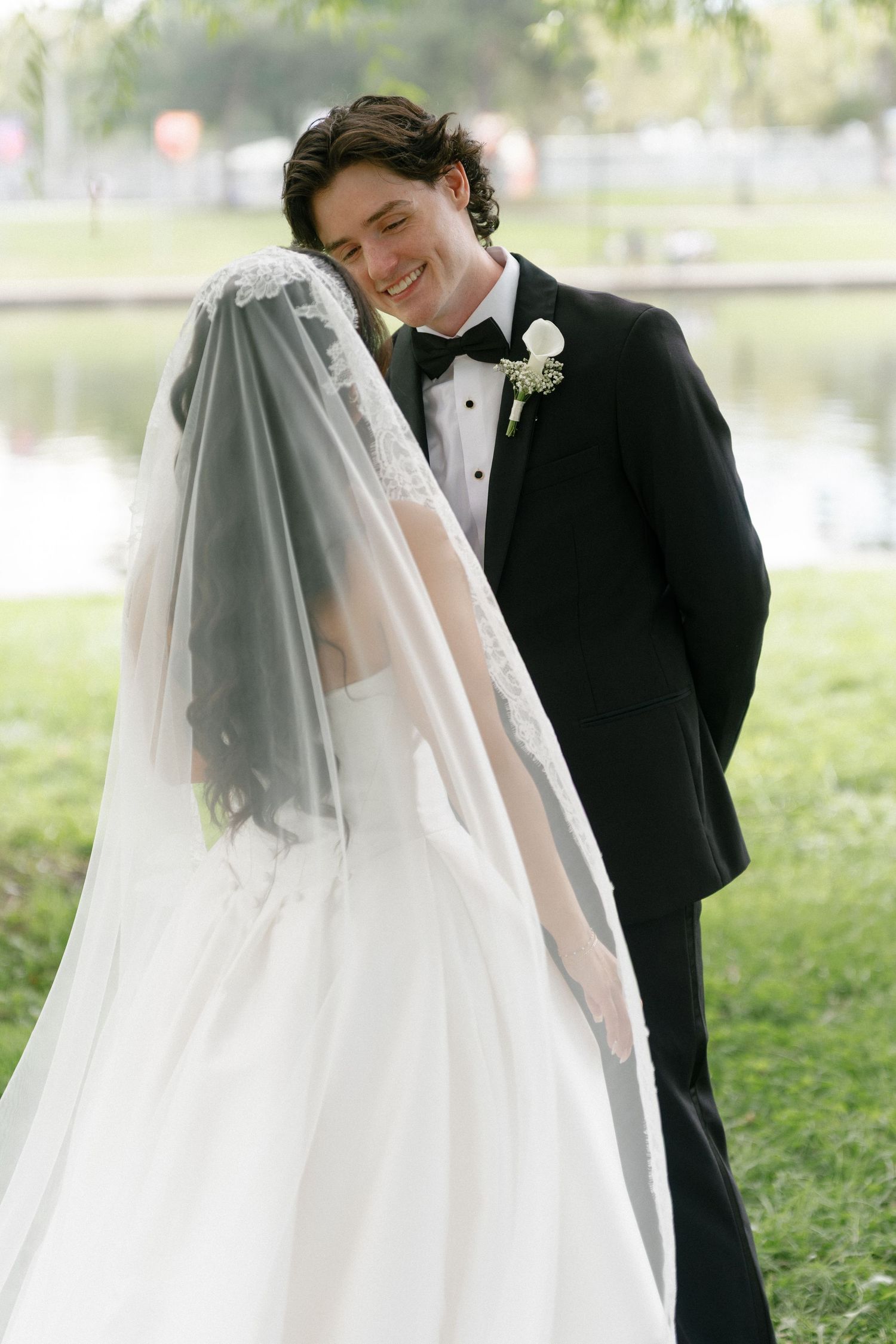 Smiling groom in black tuxedo leans toward veiled bride in white ball gown outdoors near a pond.