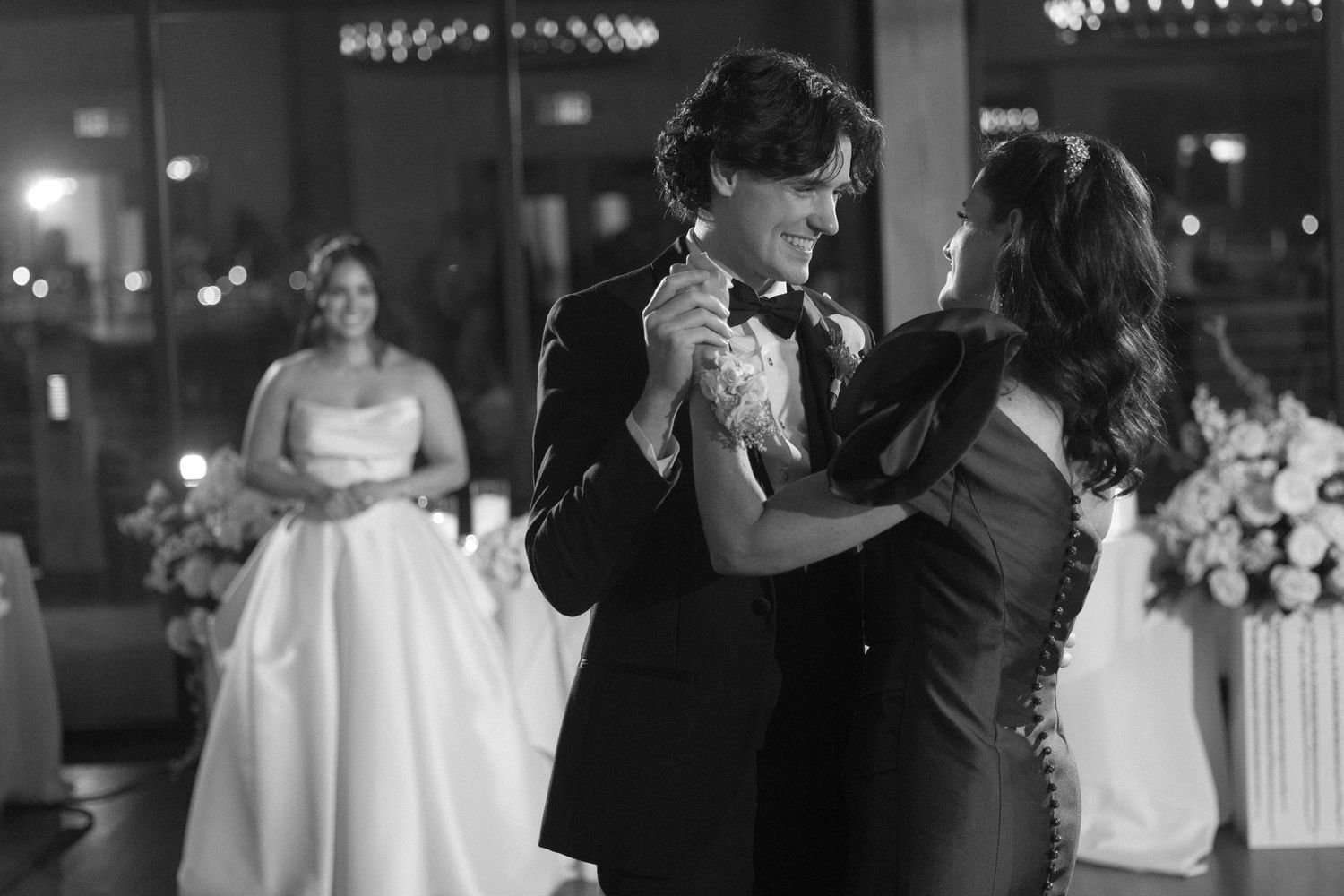 Black and white photo of a groom dancing with a woman at a wedding reception, bride visible in background.