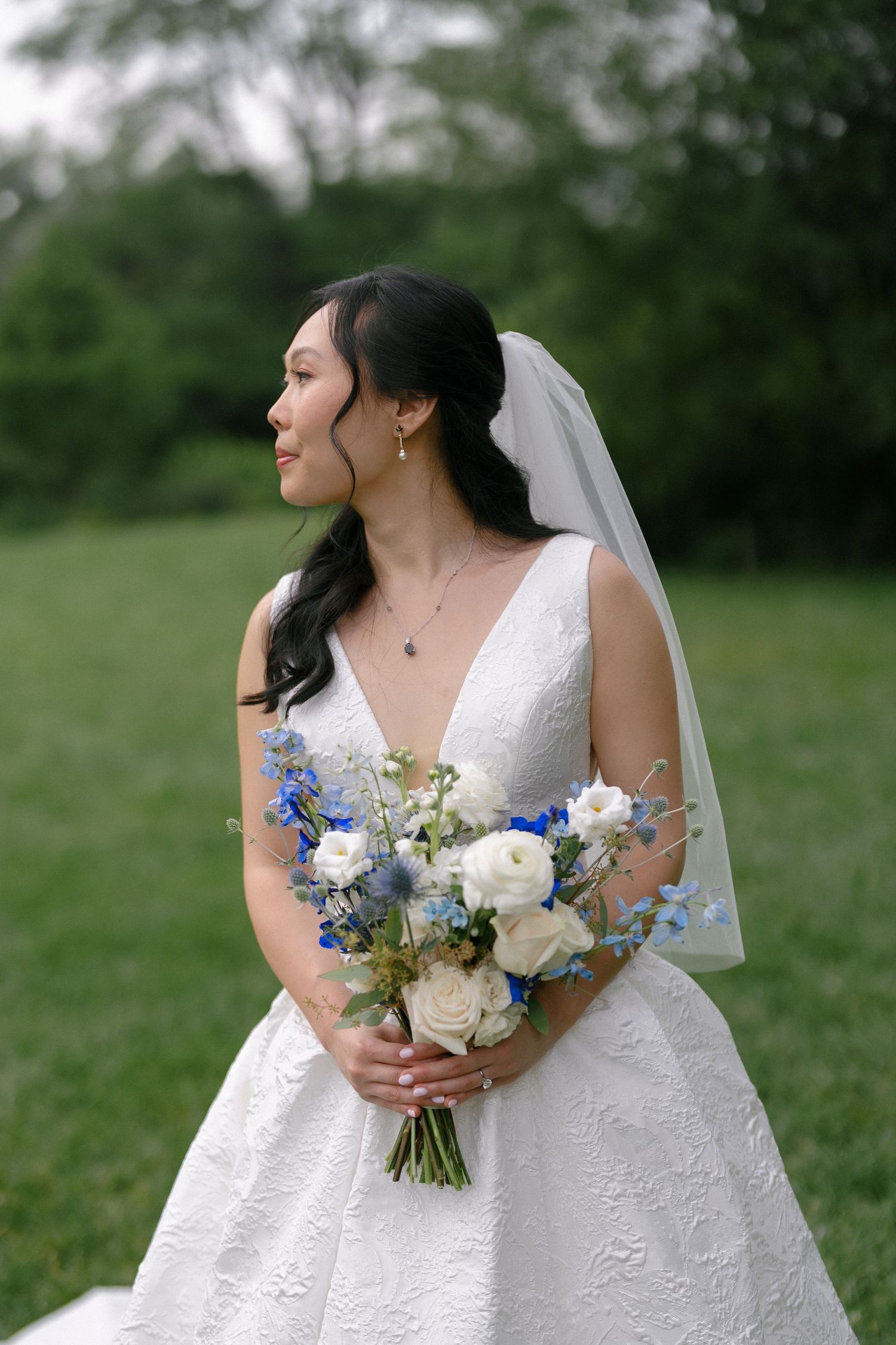 Beautiful Asian bride in lace V-neck wedding gown holds blue and white floral bouquet outdoors.