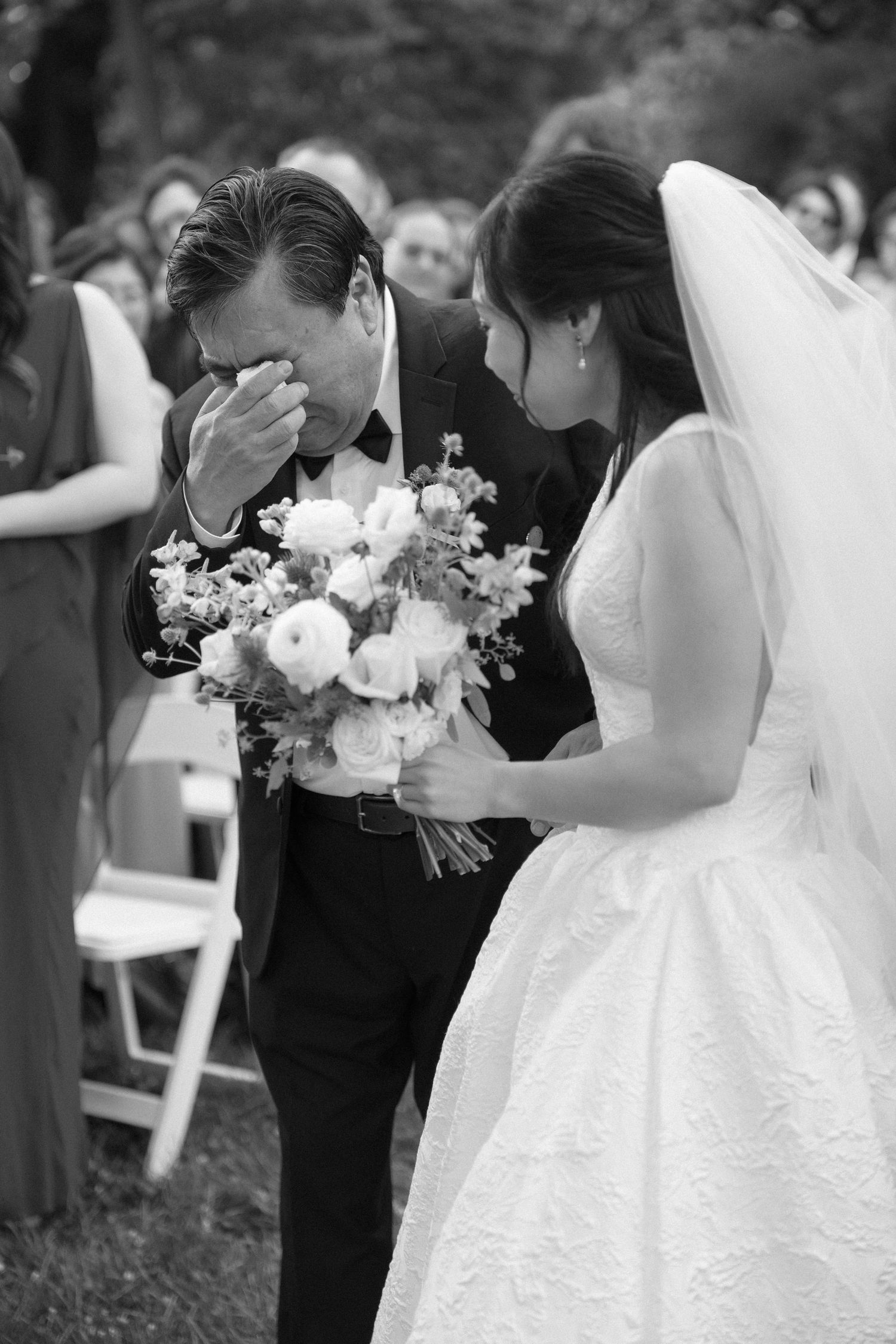 Black and white photo of emotional father wiping tears walking bride down the aisle at outdoor wedding ceremony.