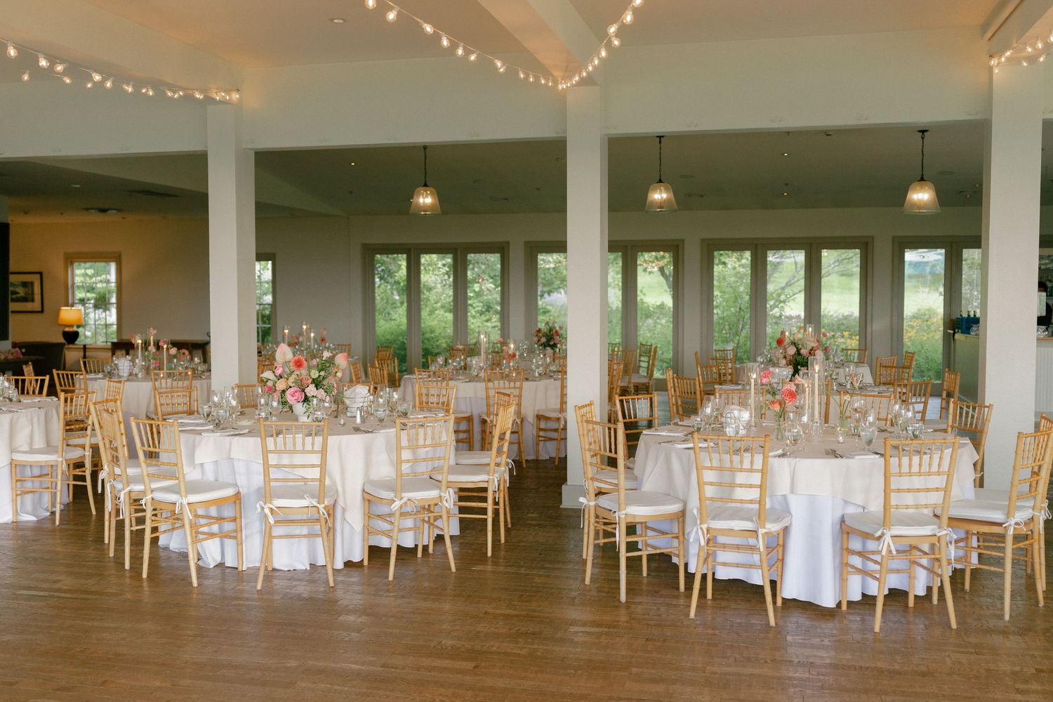 Elegant wedding reception hall with round tables, gold chiavari chairs, and string lights overhead.