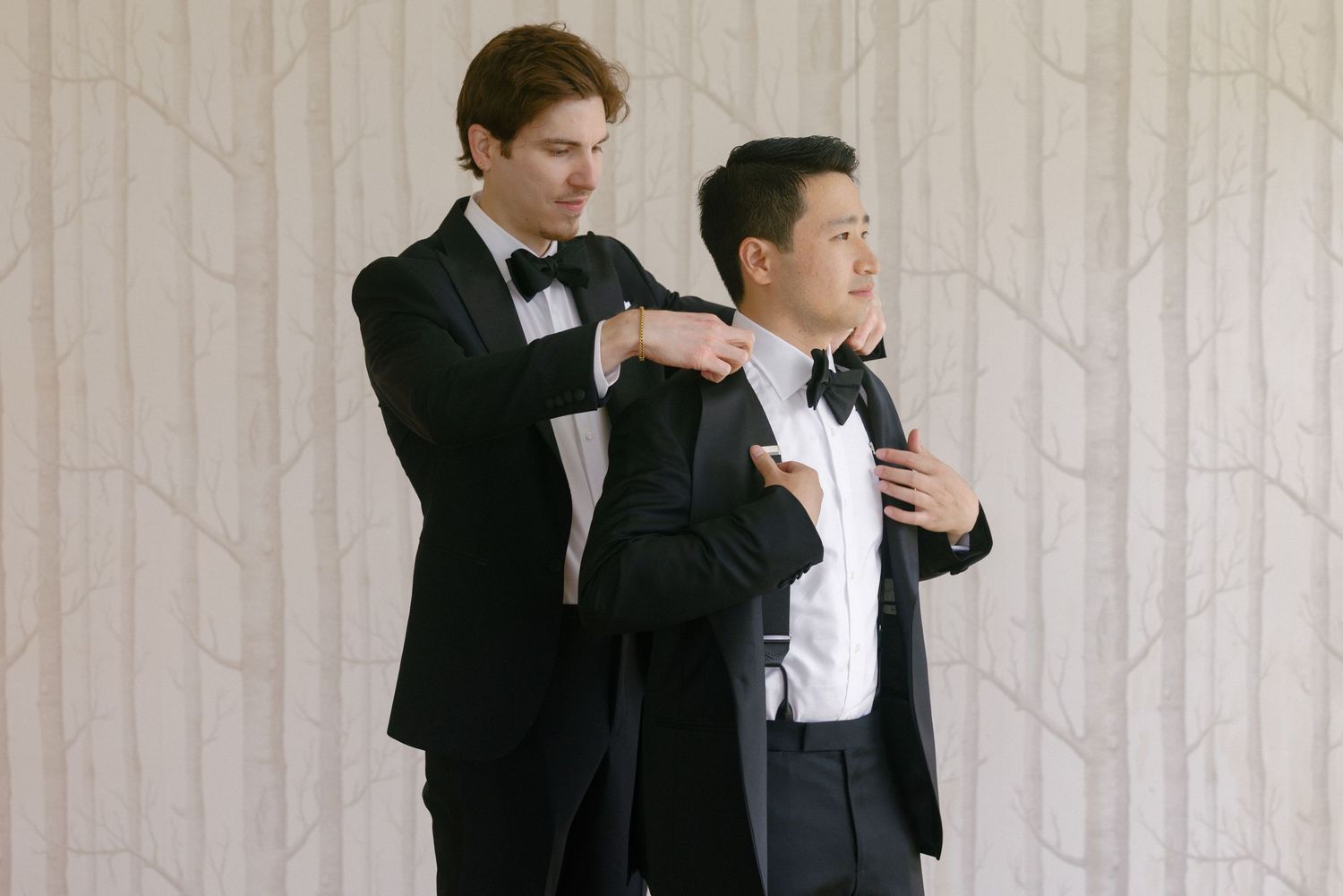 Two men in black tuxedos with bow ties, one adjusting the other's jacket collar before a formal event.