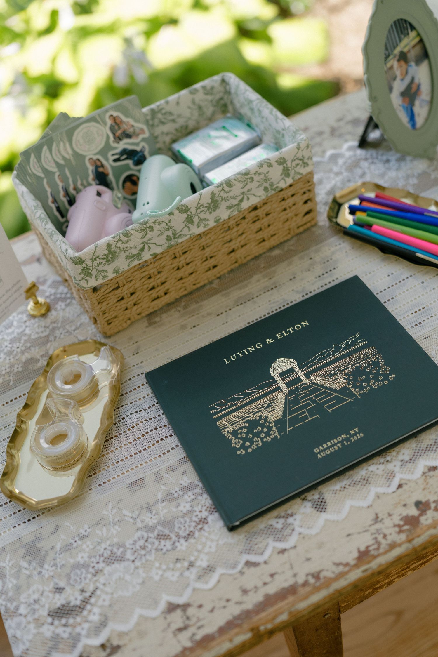 Wedding guest book table with floral basket, colorful pens, and dark green personalized couple's program book on lace tablecloth.