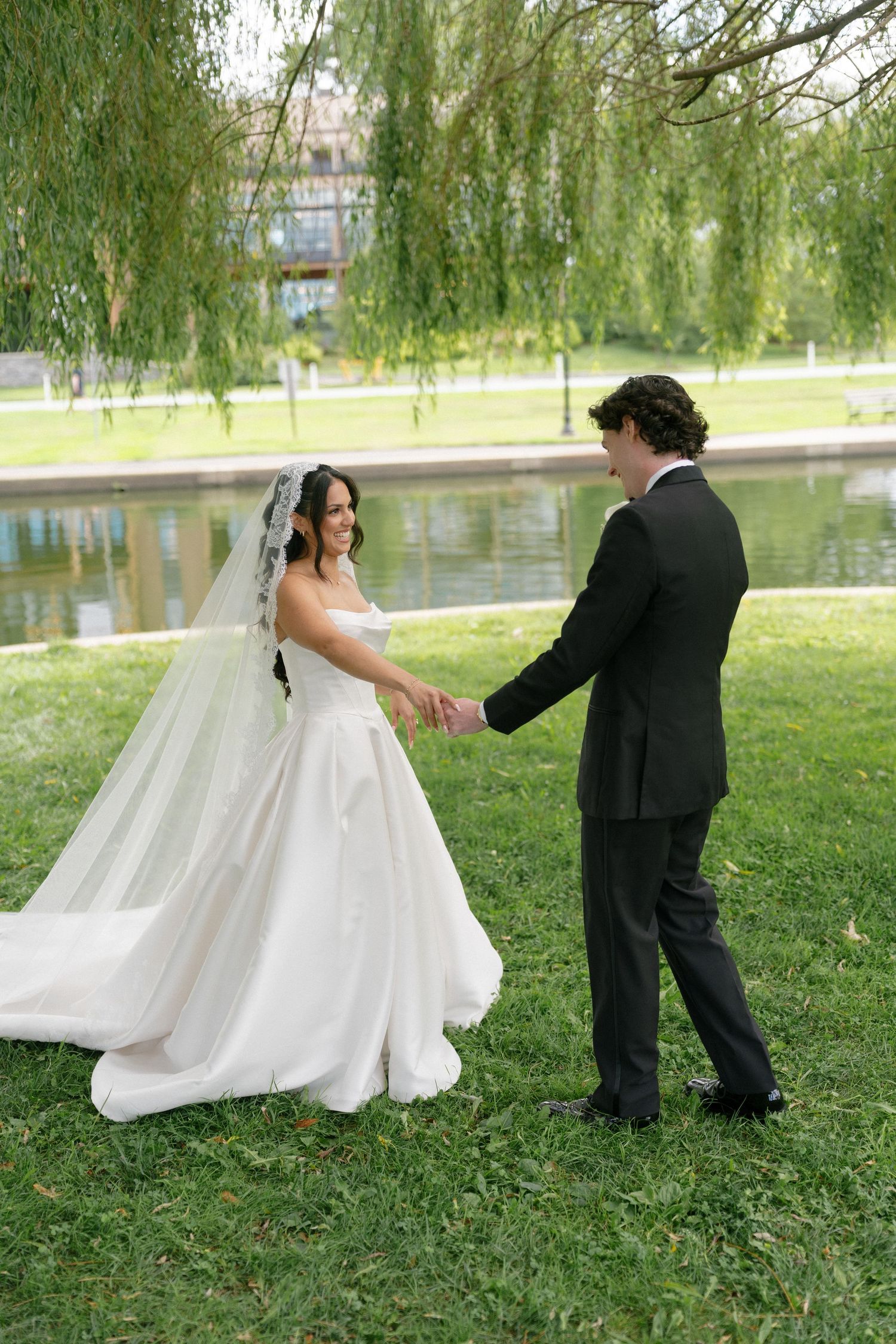 Bride and groom holding hands by a pond under a weeping willow tree on their wedding day.