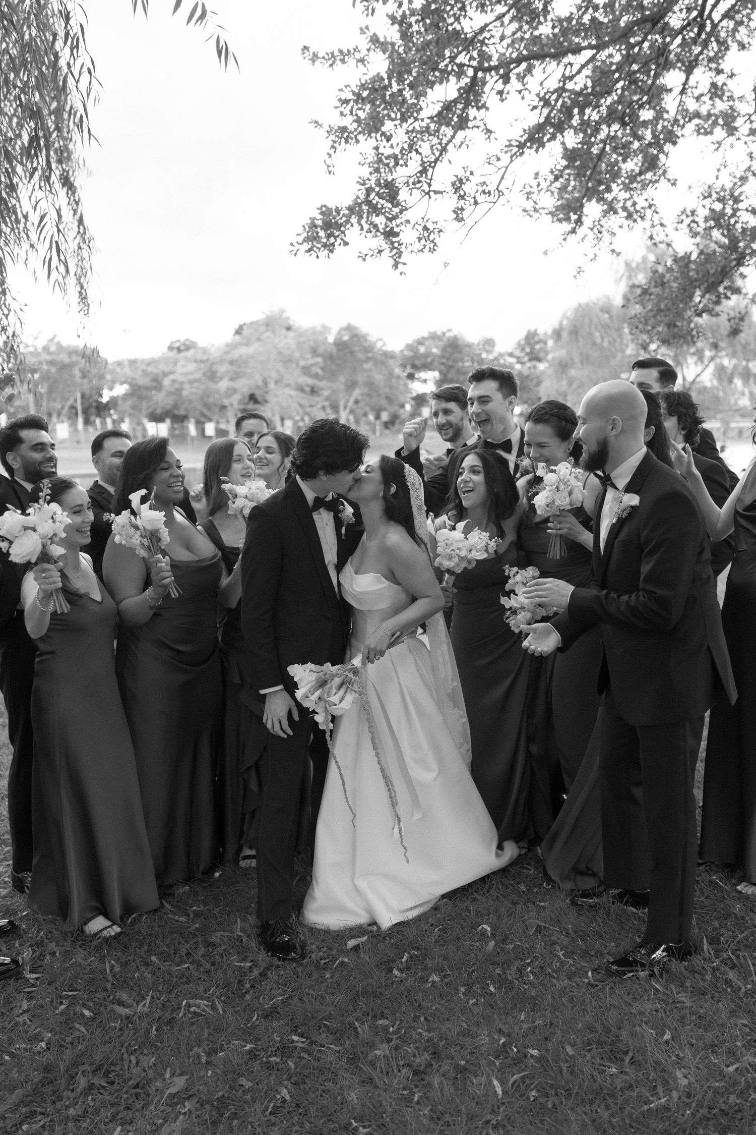 Black and white photo of bride and groom kissing surrounded by wedding party outdoors.