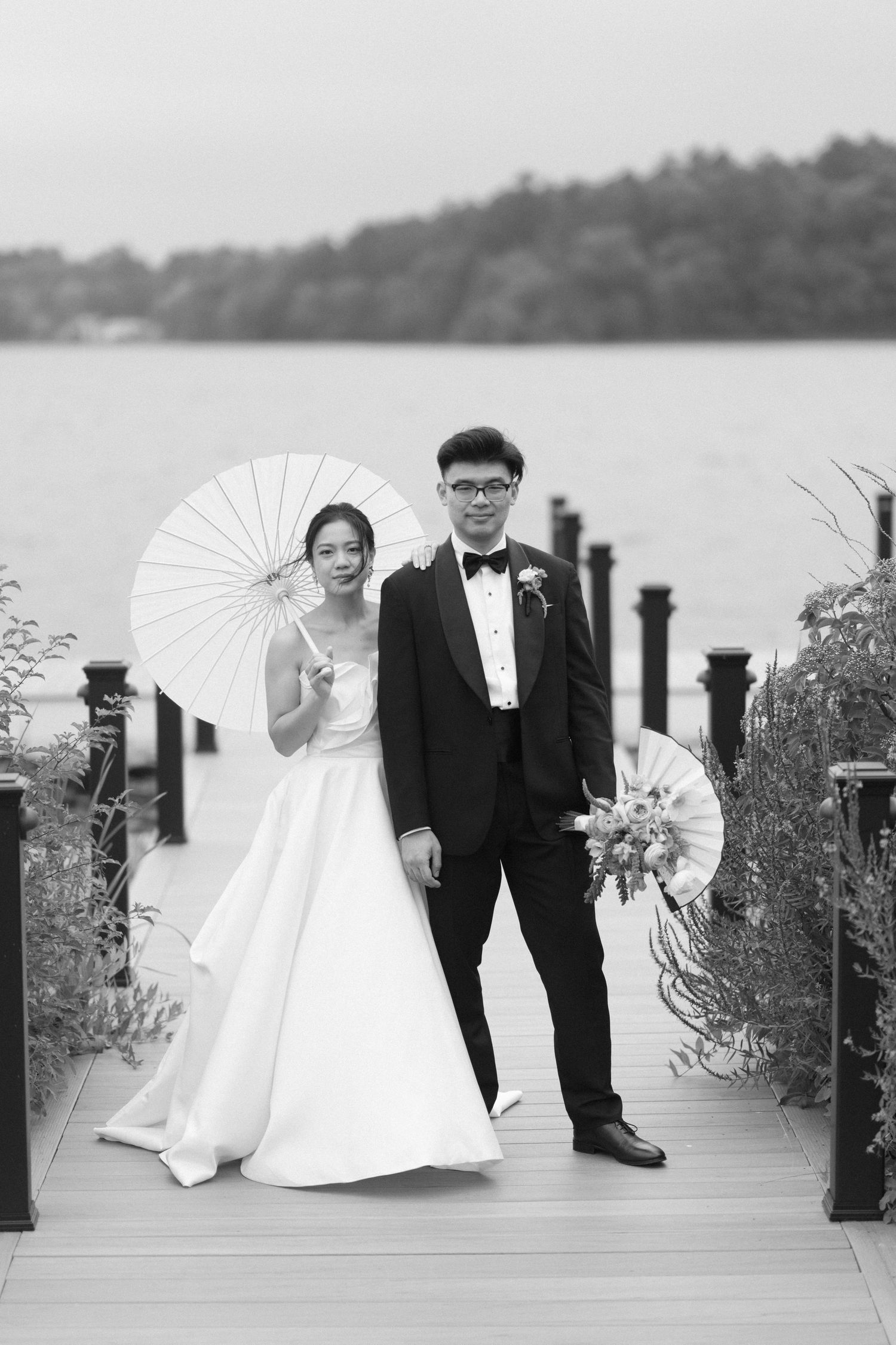 Black and white photo of Asian bride and groom posing on a lakeside dock, bride holding a white parasol.