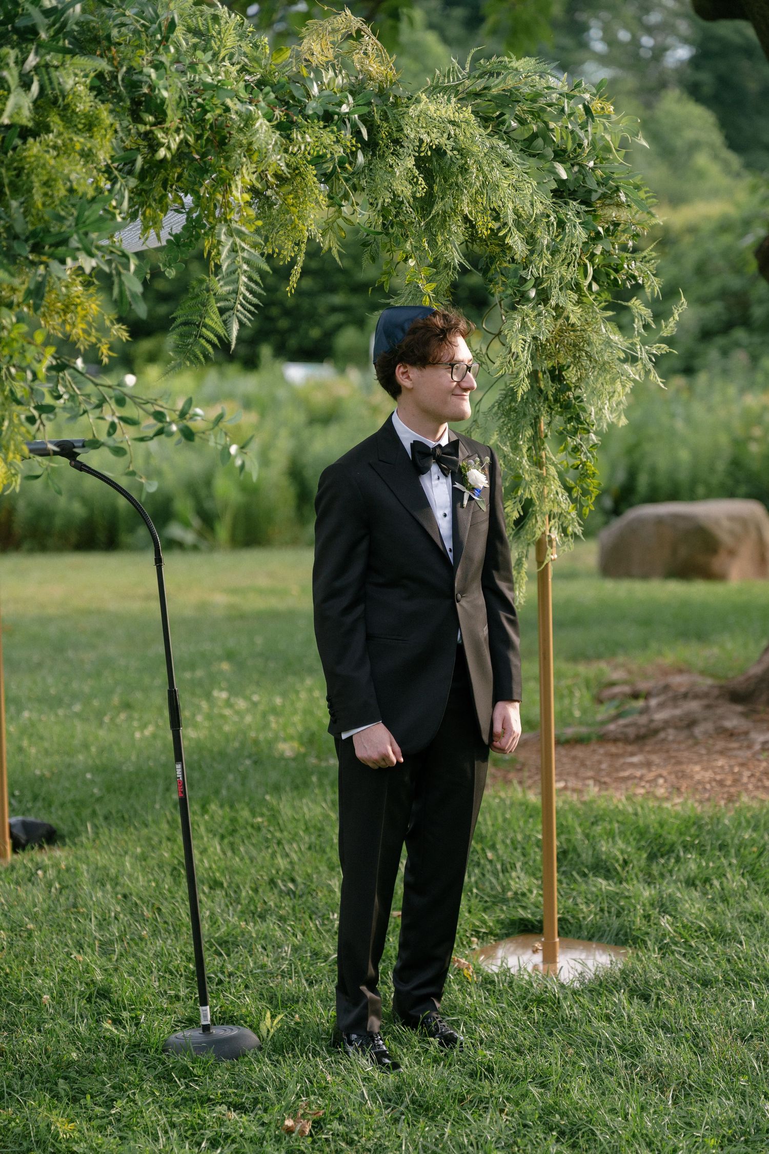 Groom in black tuxedo standing under a floral chuppah arch at an outdoor wedding ceremony.