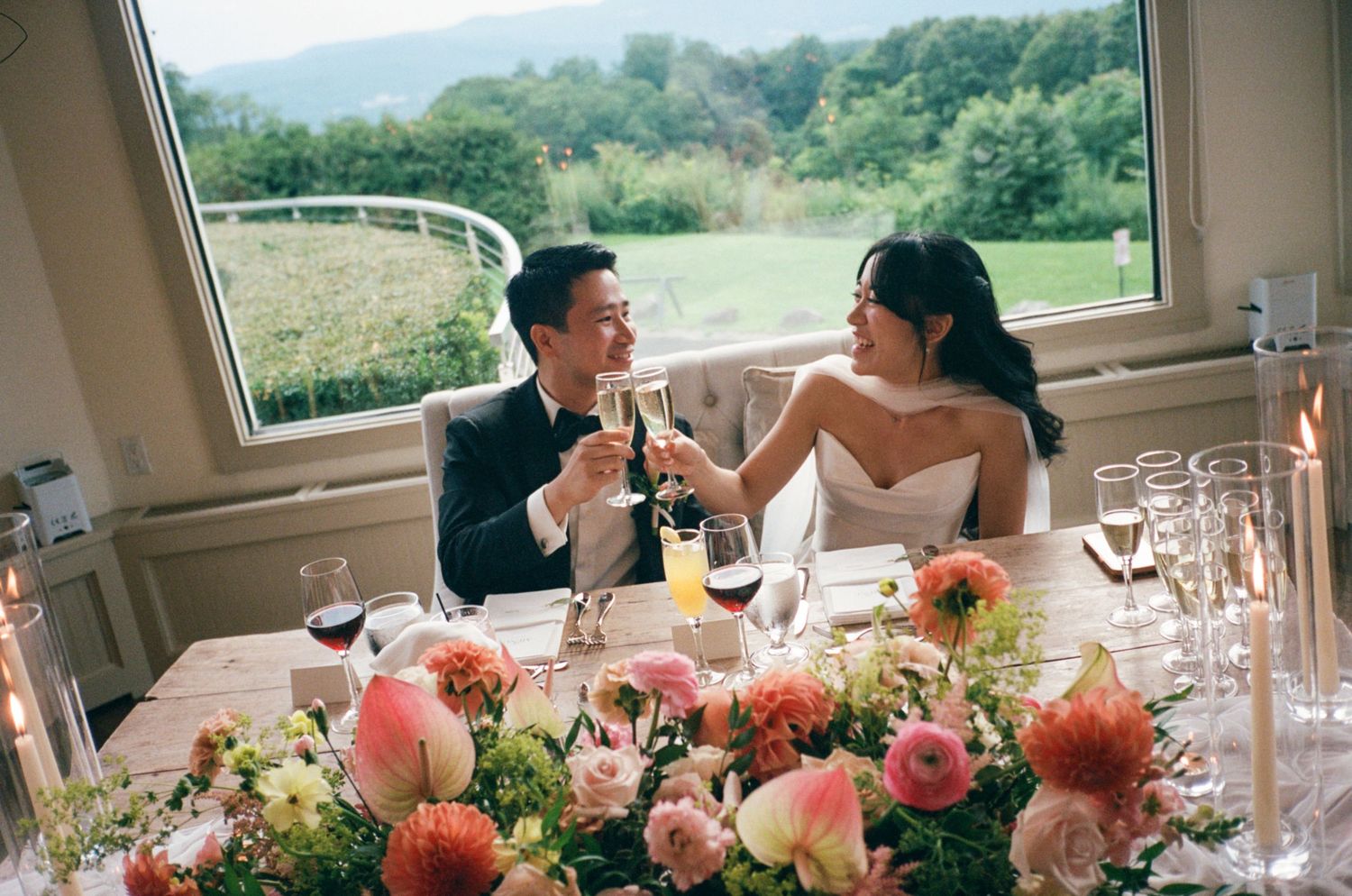 Newlywed couple toasting champagne at wedding reception table with pink floral centerpiece and scenic green landscape window view.