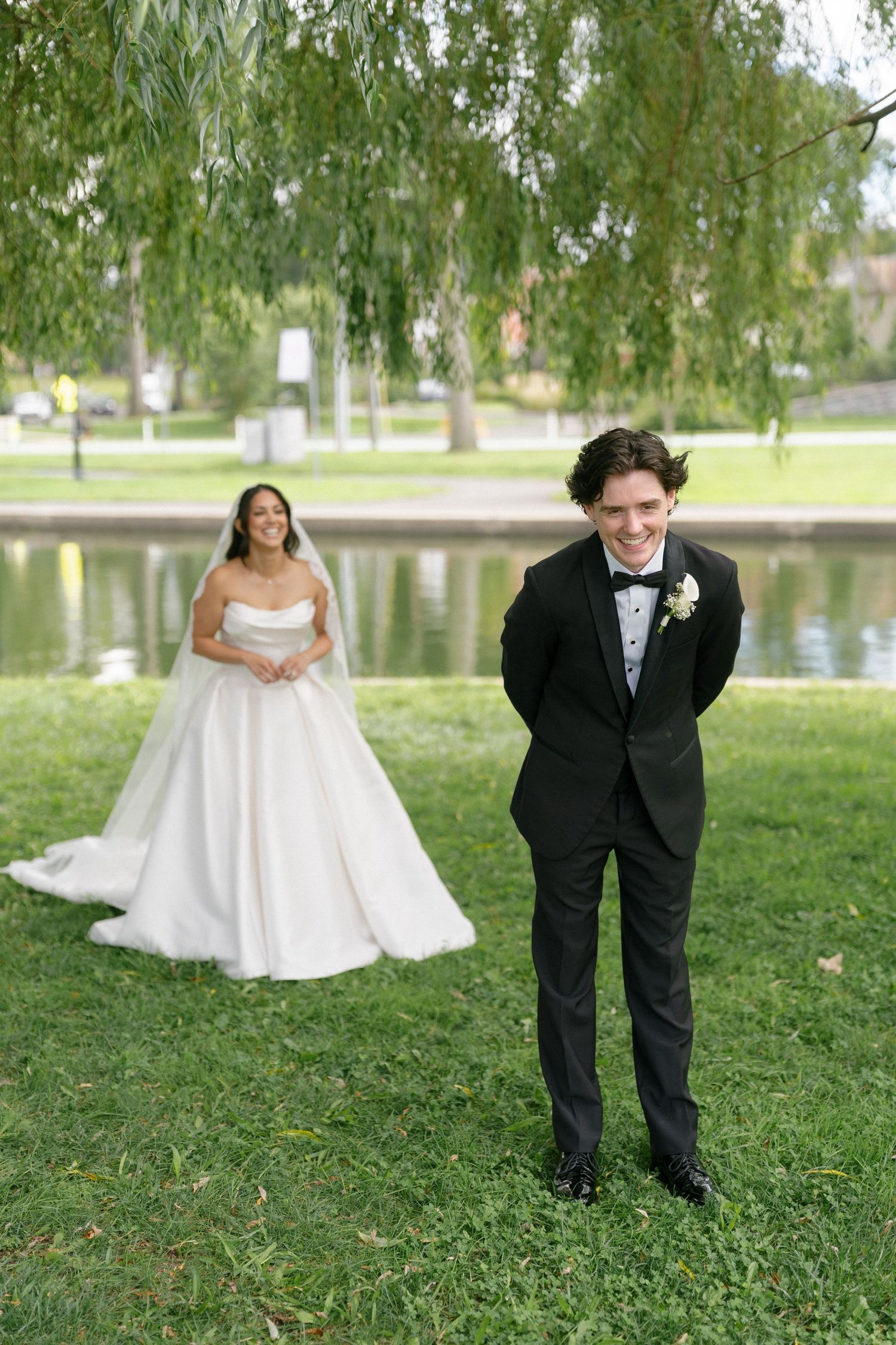 Groom in black tuxedo smiles as bride in white ball gown stands behind him near a pond.
