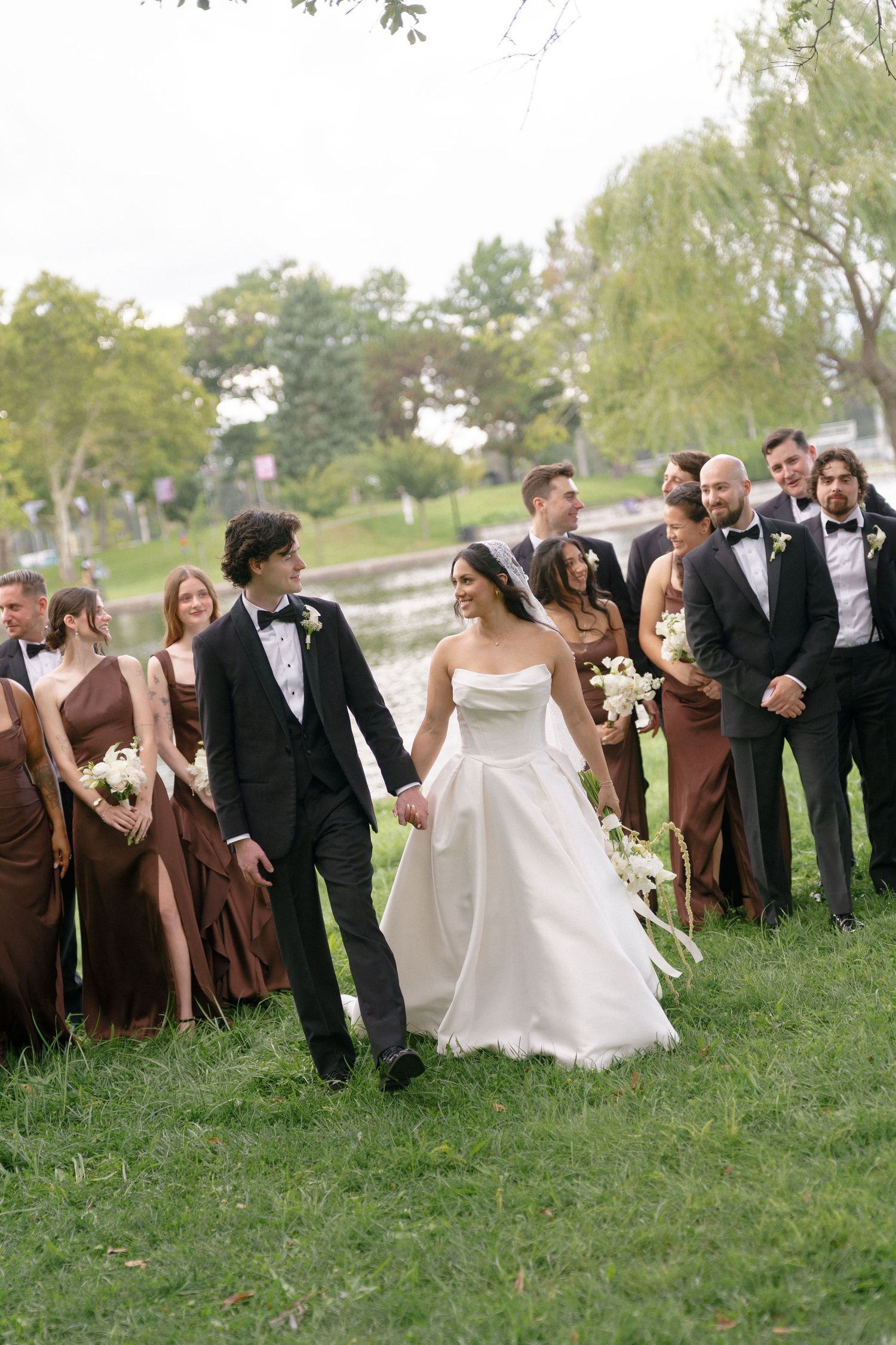 Bride and groom walking with wedding party in brown dresses and black tuxedos by a pond.