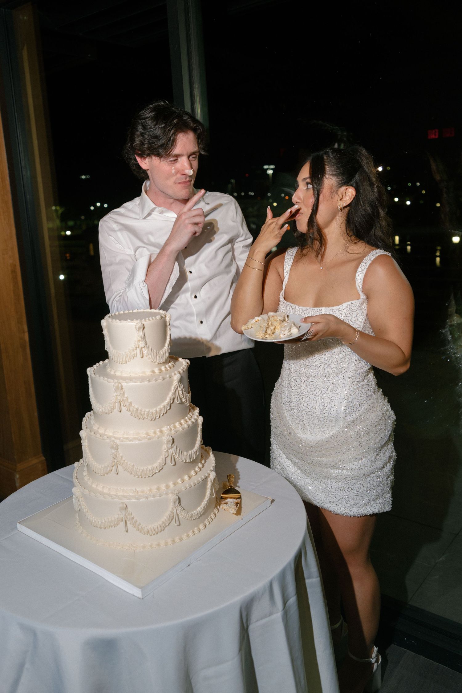 Bride and groom share wedding cake at night reception, bride wearing sparkly white mini dress.