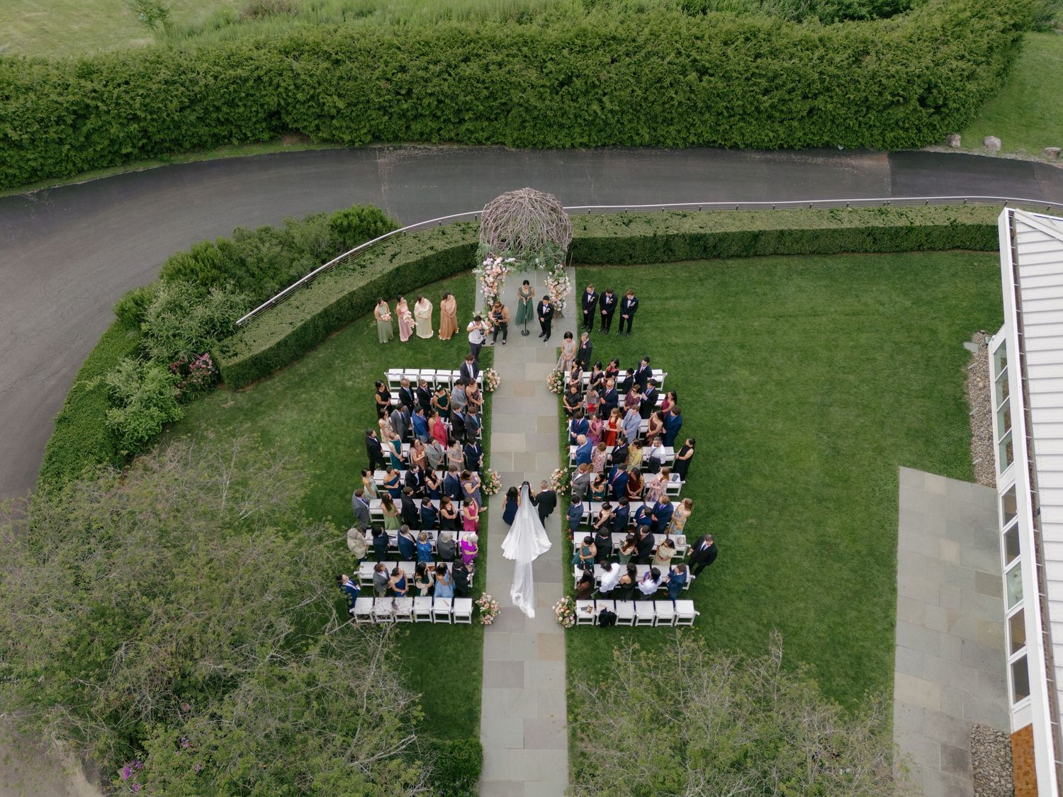 Aerial drone view of an outdoor wedding ceremony with guests seated in white chairs on a green lawn.