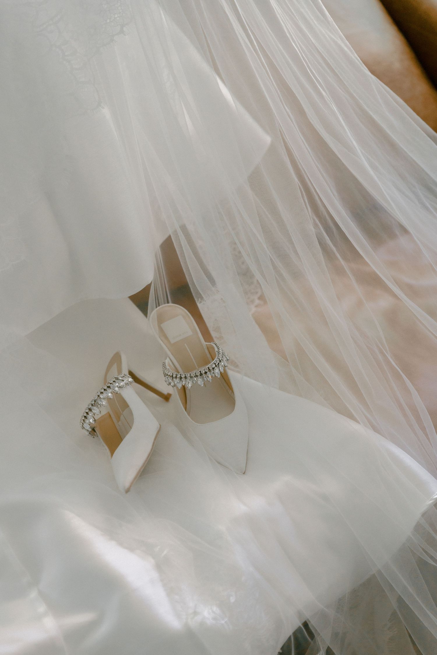 White pointed-toe bridal heels with crystal ankle strap detail resting on flowing tulle wedding veil.