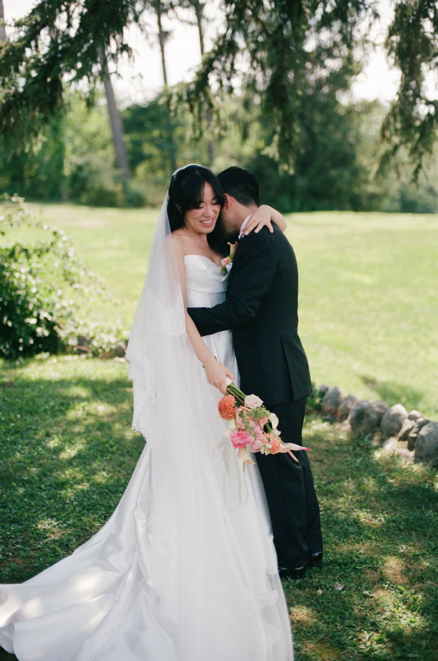 Bride and groom embracing outdoors, she wears a white gown with veil, holding a pink bouquet.