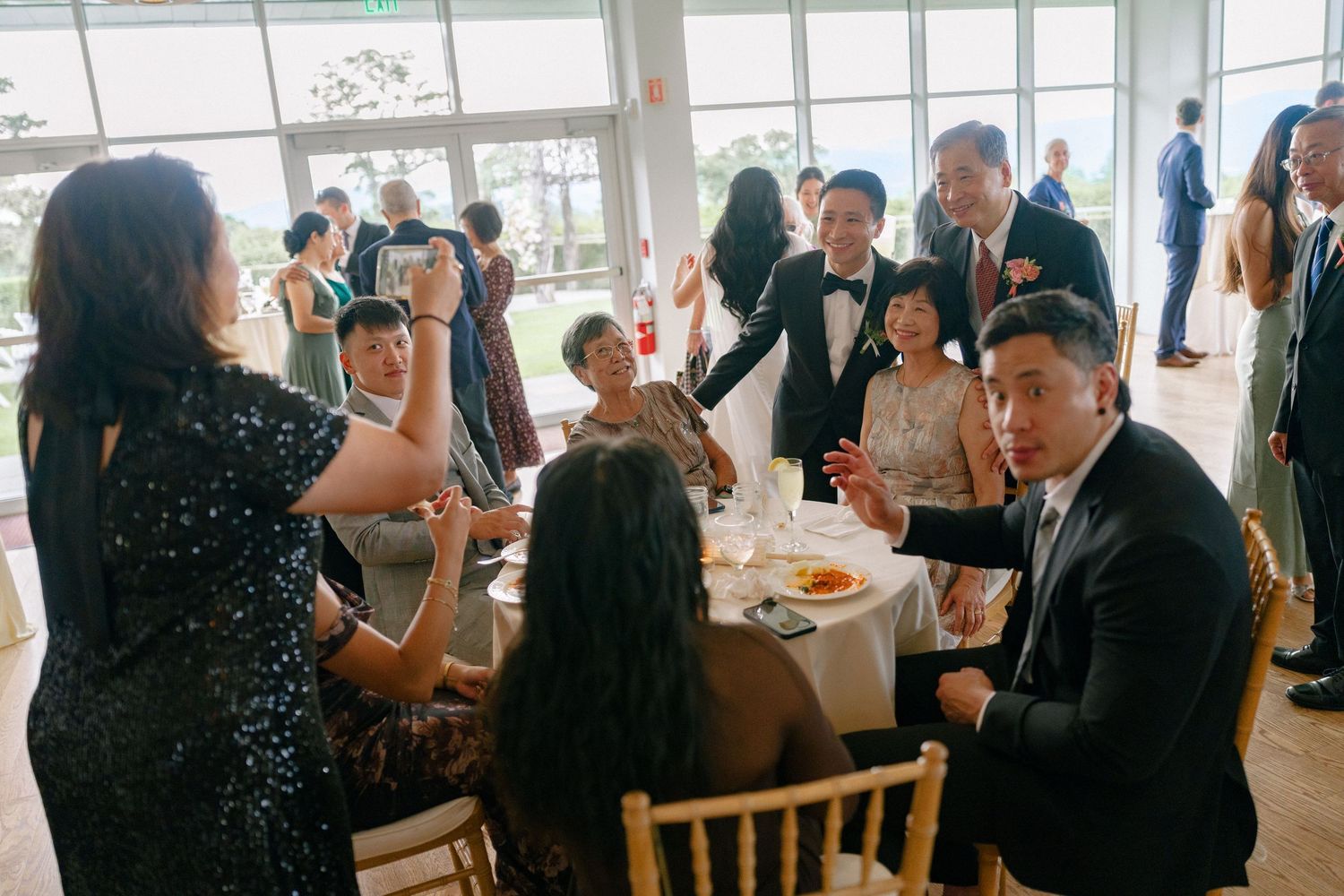 Wedding reception guests mingling and toasting at elegantly set round tables in bright venue.