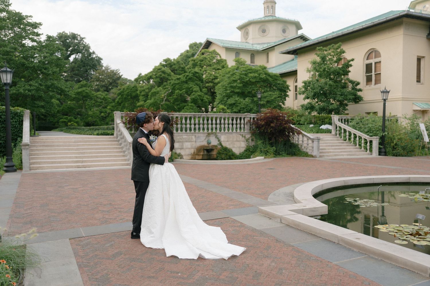 Bride and groom share a romantic kiss at an elegant outdoor wedding venue with stone architecture and lush greenery.